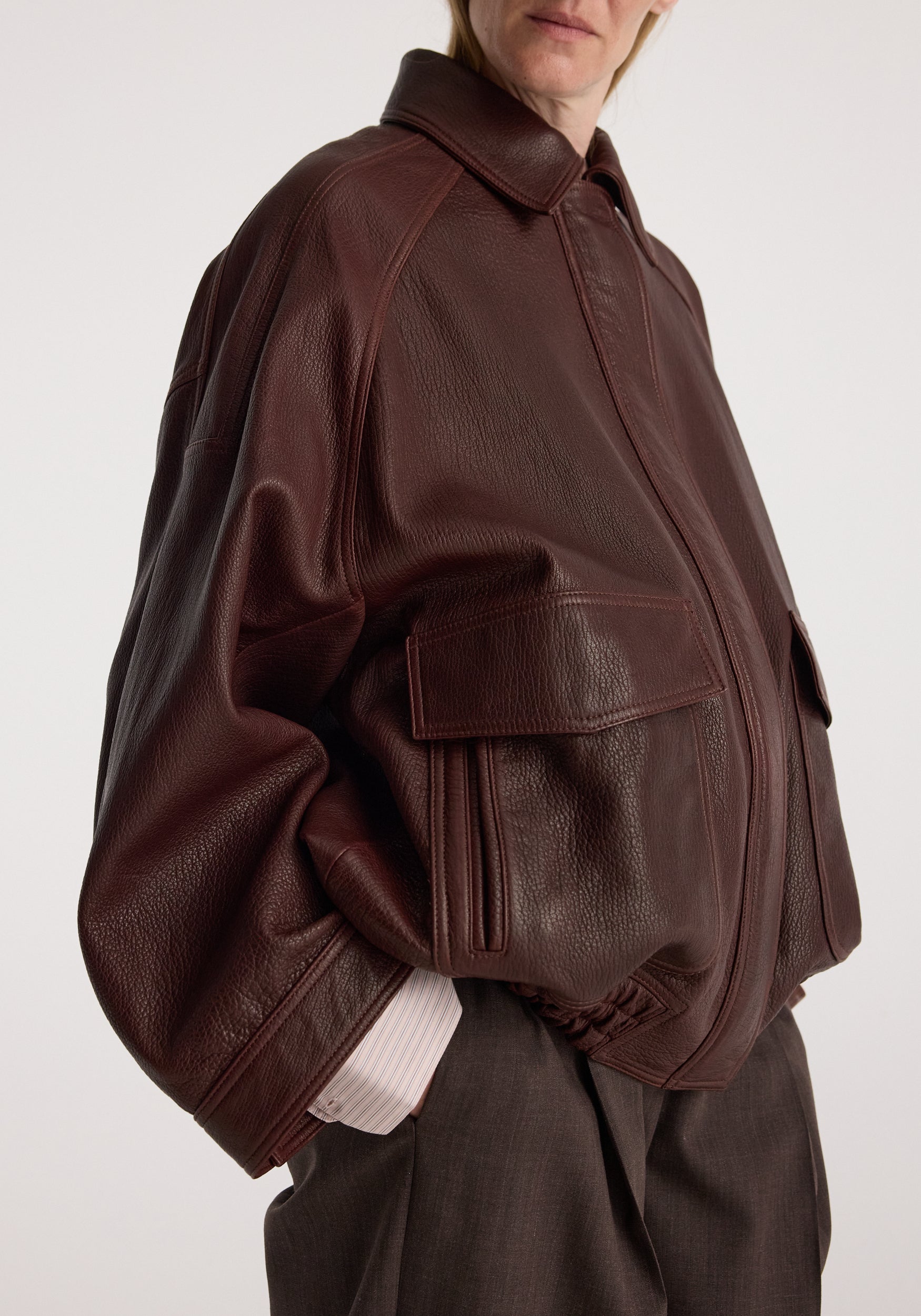 Close-up of a person wearing a chestnut Grain leather aviator jacket with large front pockets and a spread collar, styled with dark pants and a light shirt cuff visible.