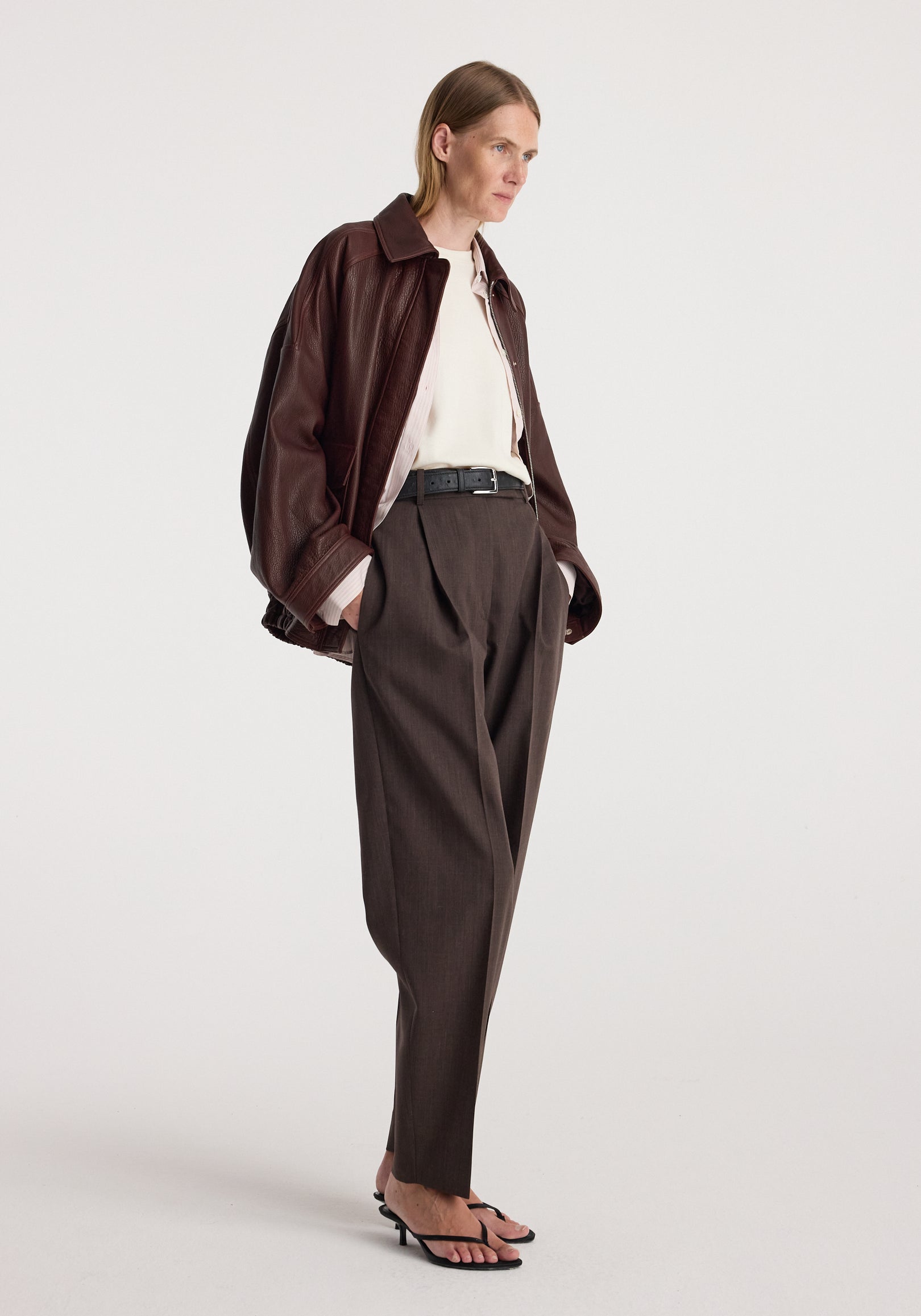 Woman wearing a chestnut grain leather aviator jacket over a white top and dark brown trousers, styled with black sandals and a black belt.