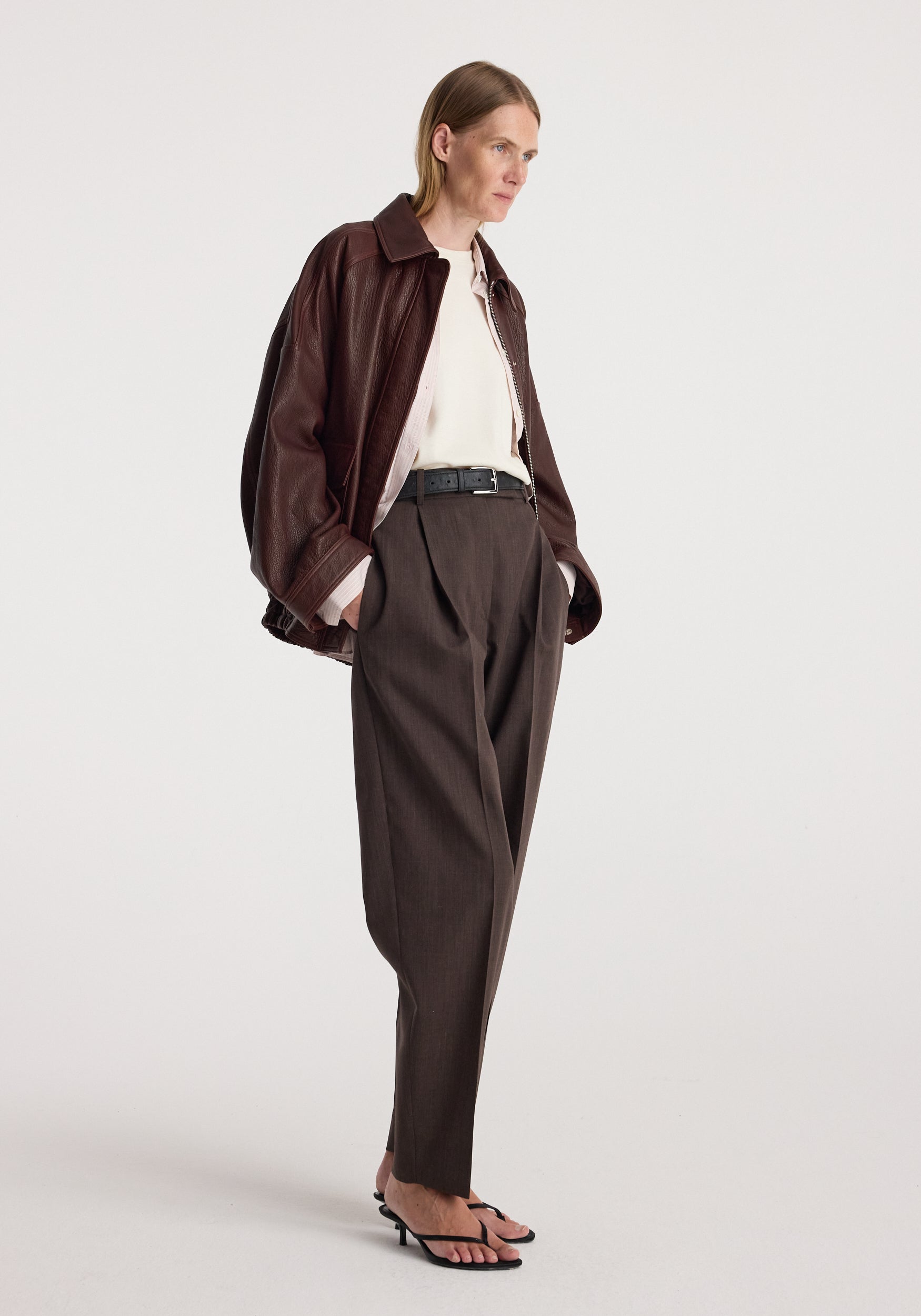 Woman wearing a chestnut grain leather aviator jacket over a white top and dark brown trousers, styled with black sandals and a black belt.
