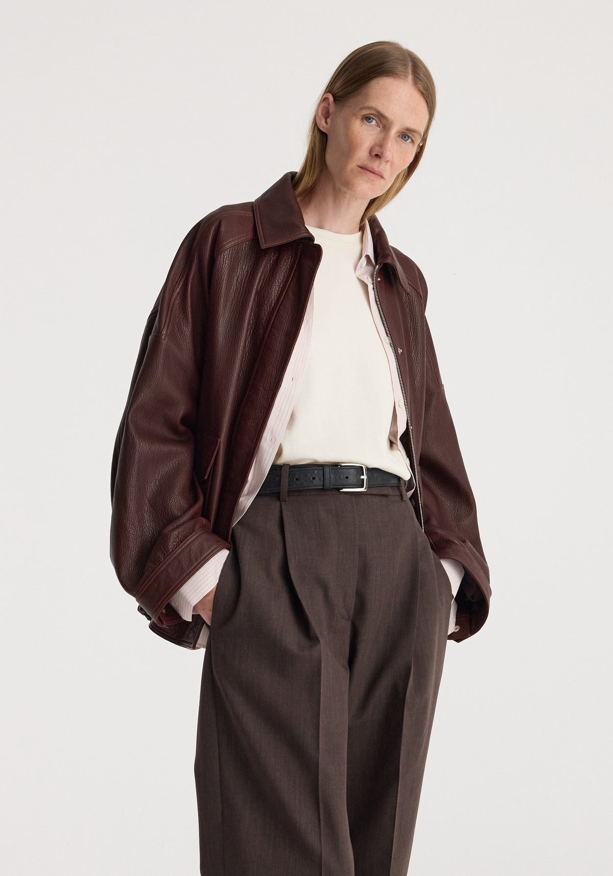 Woman wearing a chestnut grain leather aviator jacket over a white top and brown wide-leg trousers with hands in pockets against a plain background