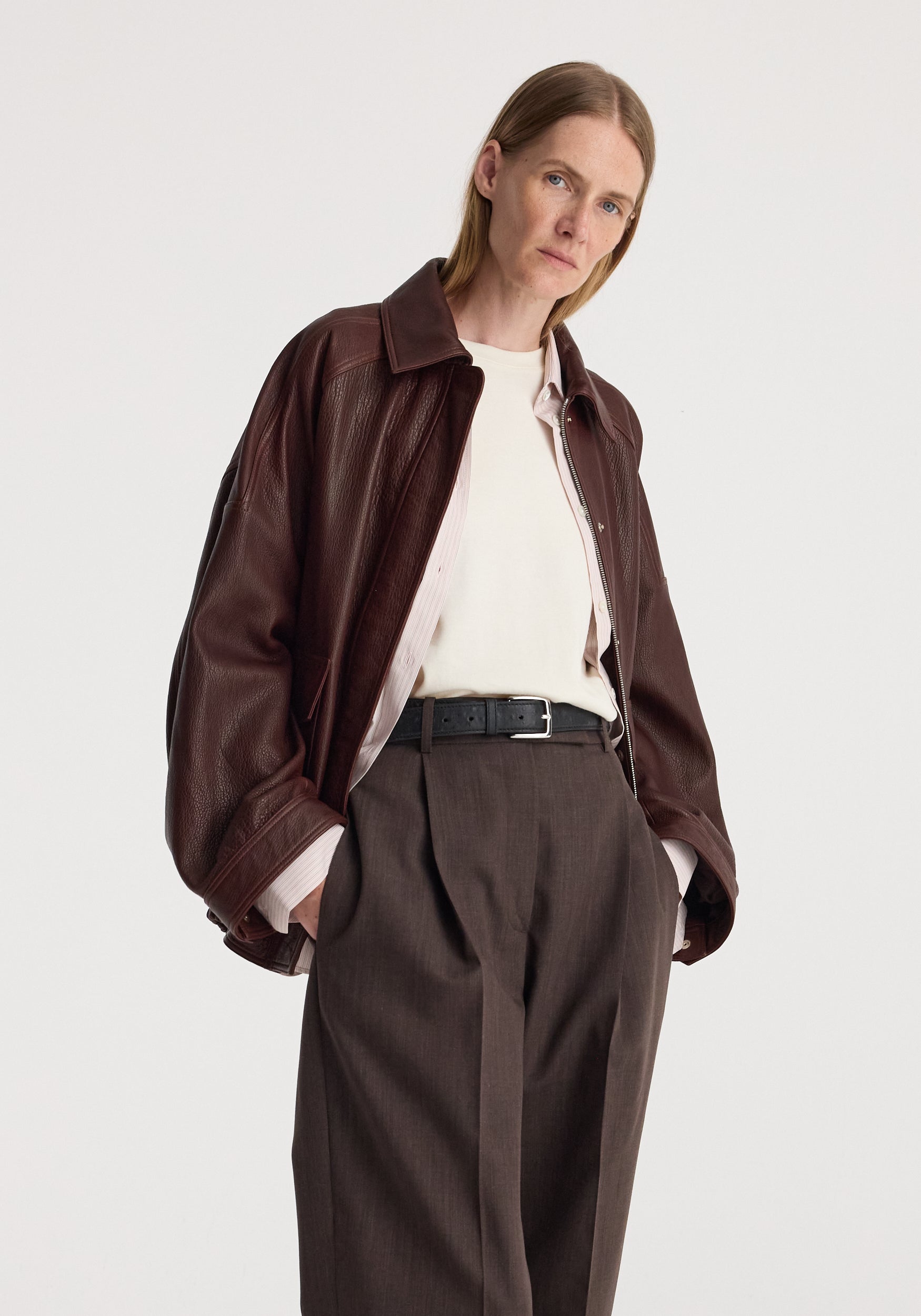Woman wearing a chestnut grain leather aviator jacket over a white top and brown wide-leg trousers with hands in pockets against a plain background