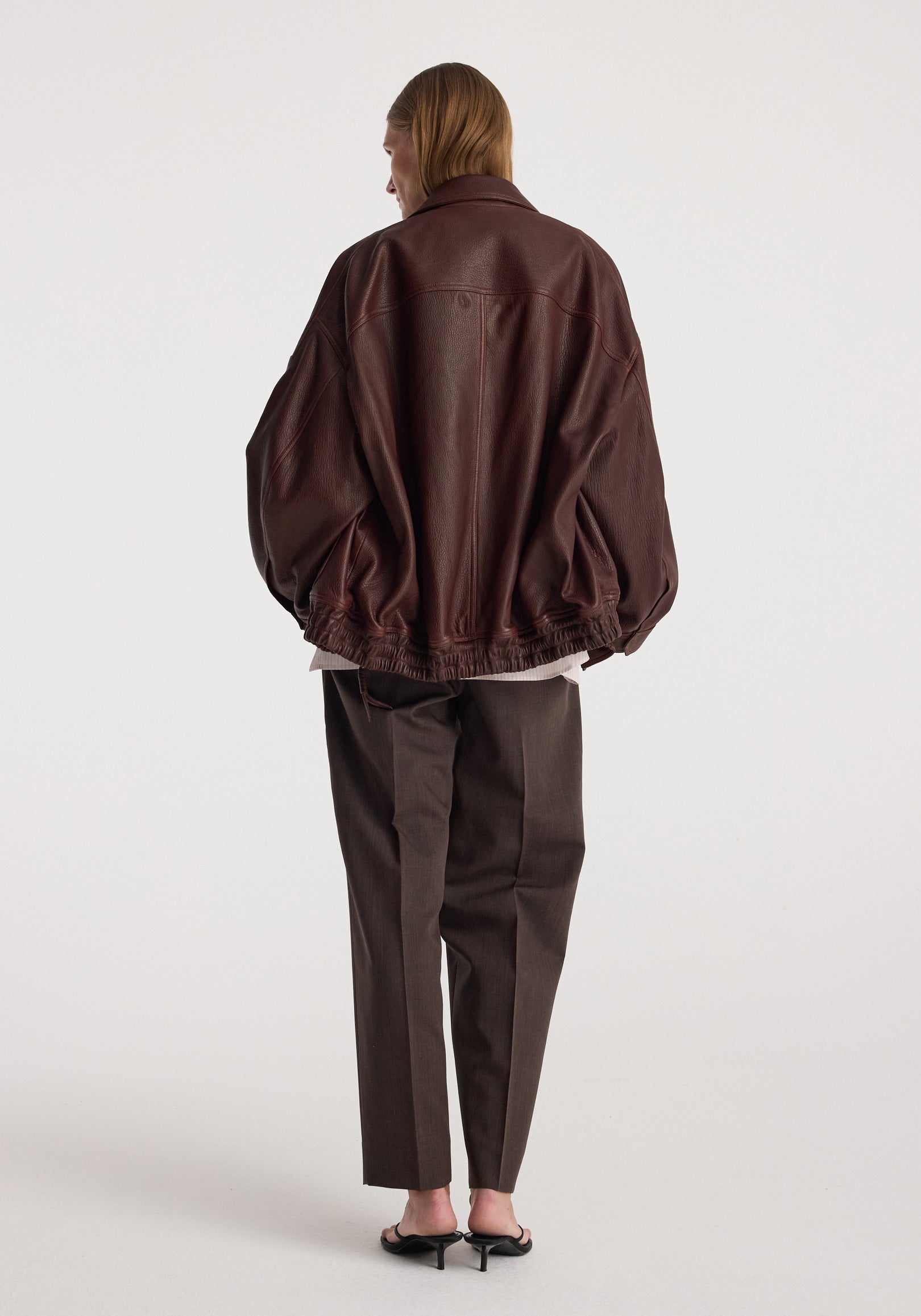 Back view of a person wearing a loose-fitting Grain leather aviator jacket | chestnut with dark tailored pants and black heels against a plain background.