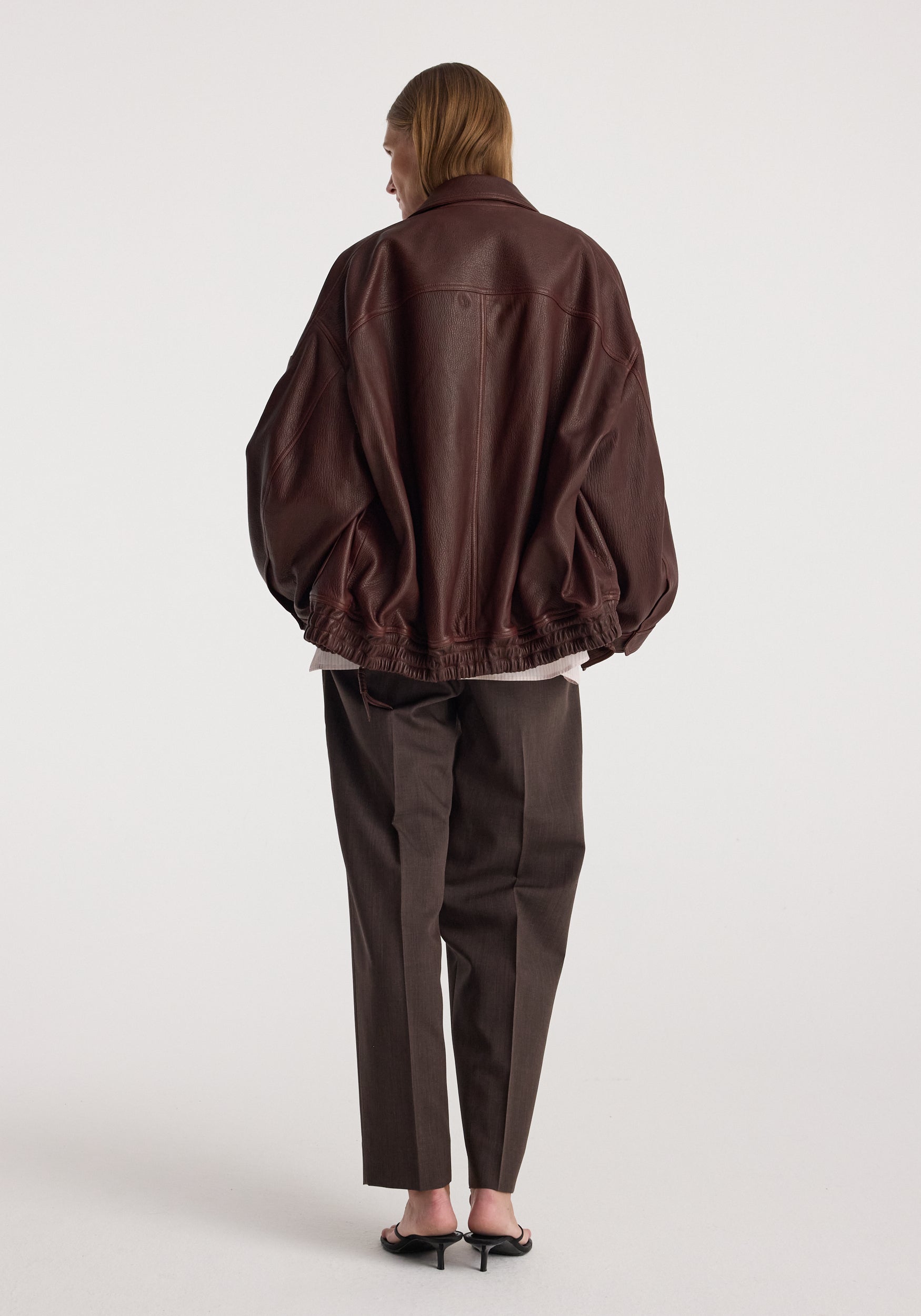 Back view of a person wearing a loose-fitting Grain leather aviator jacket | chestnut with dark tailored pants and black heels against a plain background.