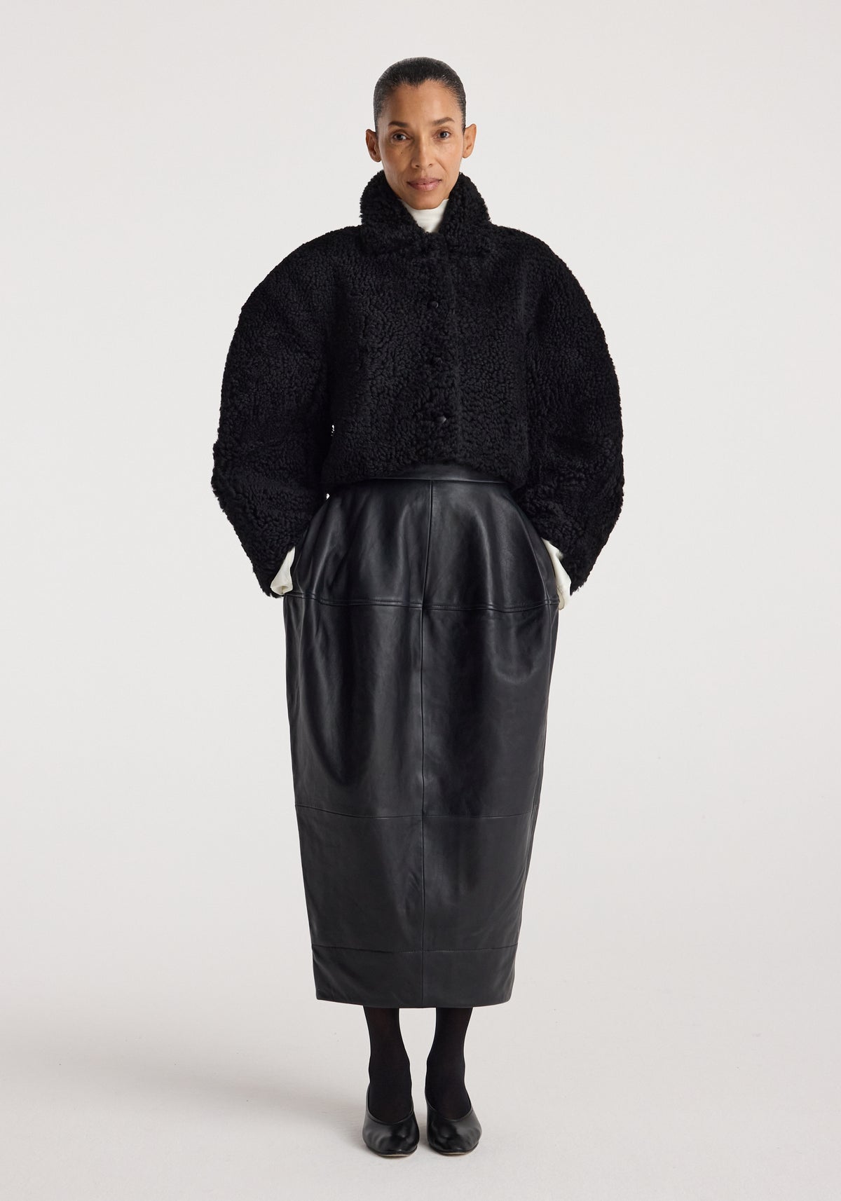 Woman wearing a black rounded shearling jacket | black with a high collar, paired with a long black leather skirt, black tights, and black shoes against a plain background.