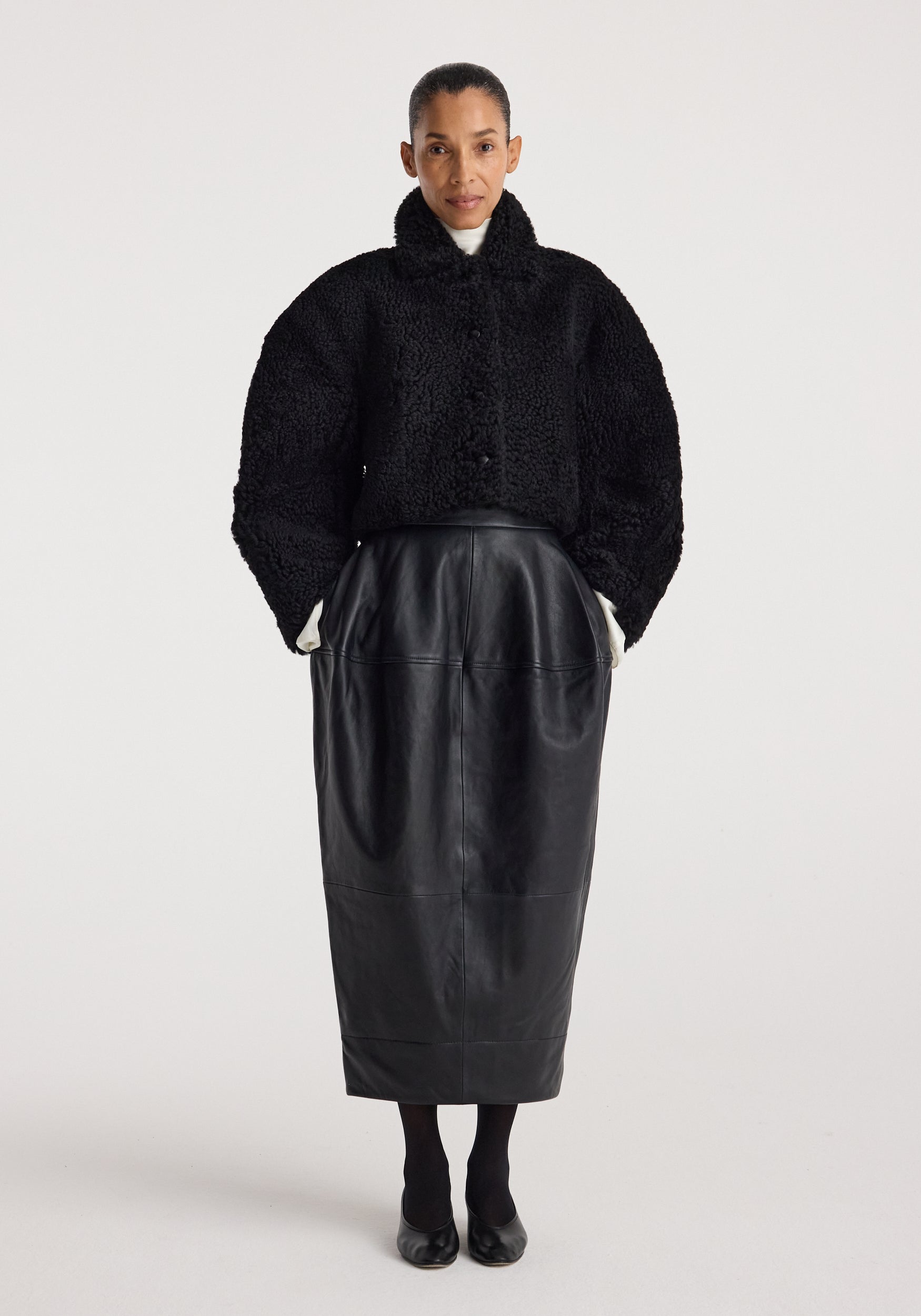 Woman wearing a black rounded shearling jacket | black with a high collar, paired with a long black leather skirt, black tights, and black shoes against a plain background.