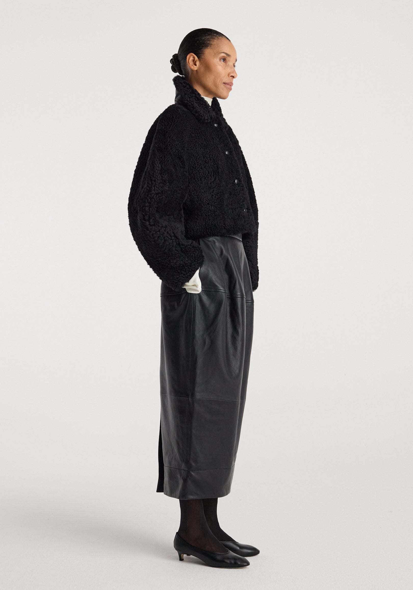 Woman wearing a black rounded shearling jacket | black with a high collar and button closure, paired with a long black leather skirt, black tights, and black low-heeled shoes.