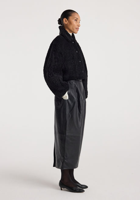 Woman wearing a black rounded shearling jacket | black with a high collar, paired with a long black leather skirt, black tights, and black shoes against a plain background.