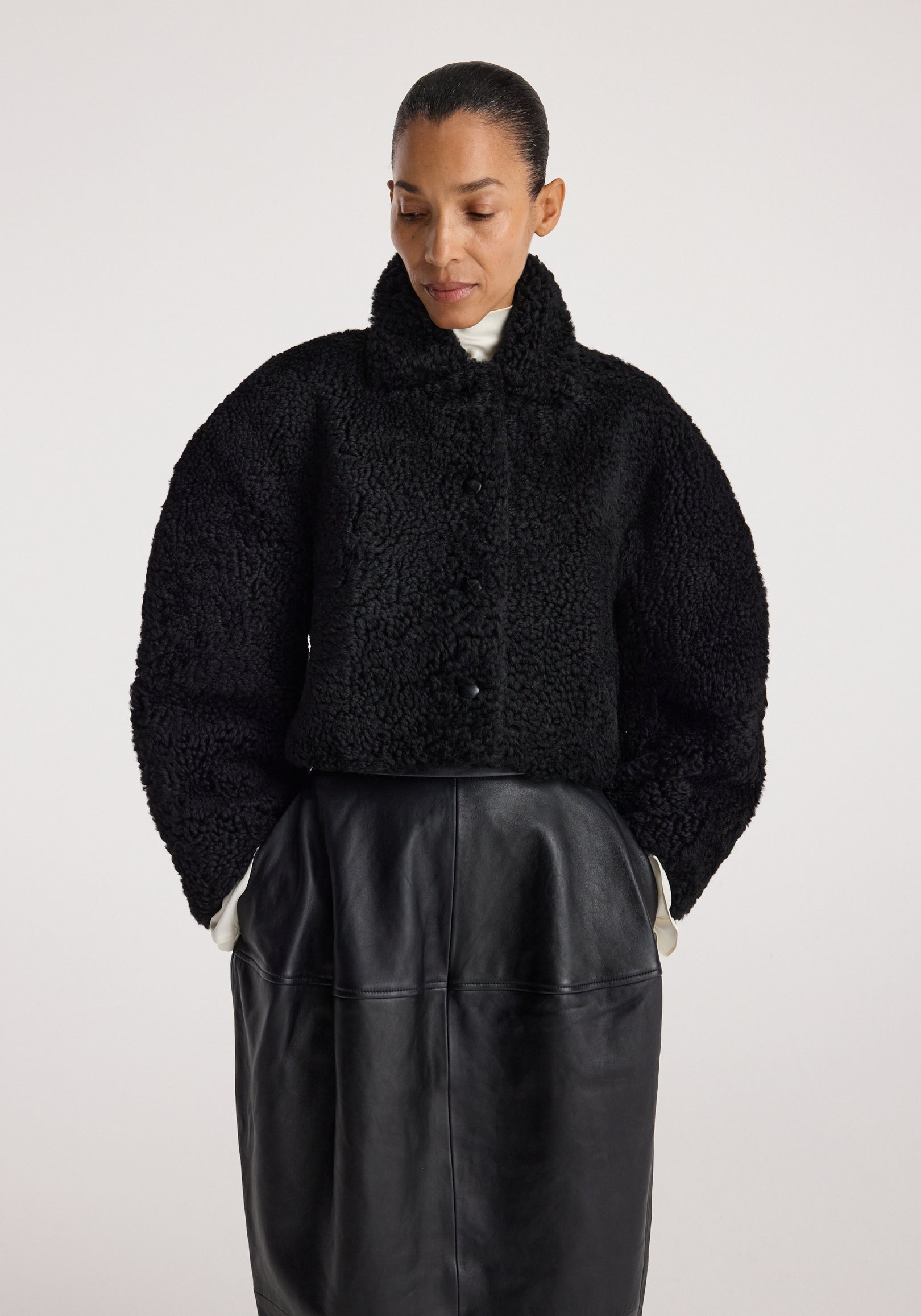 Woman wearing a black rounded shearling jacket | black with a high collar, paired with a black leather skirt, standing against a plain light background.