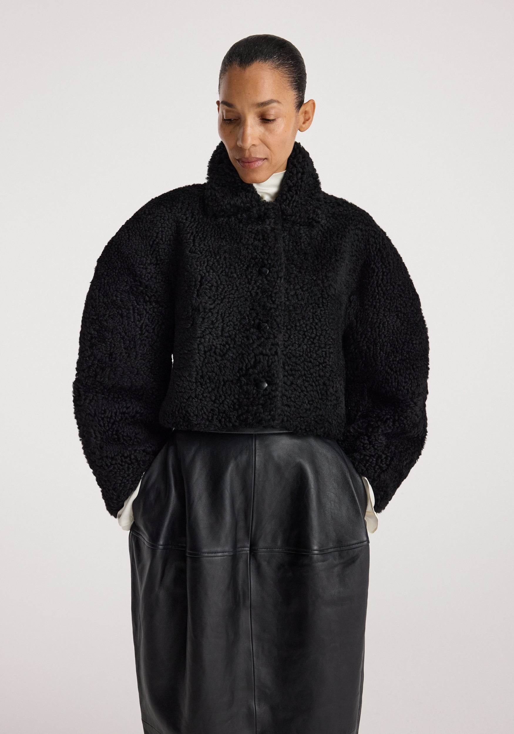 Woman wearing a black rounded shearling jacket | black with a high collar, paired with a black leather skirt, standing against a plain light background.