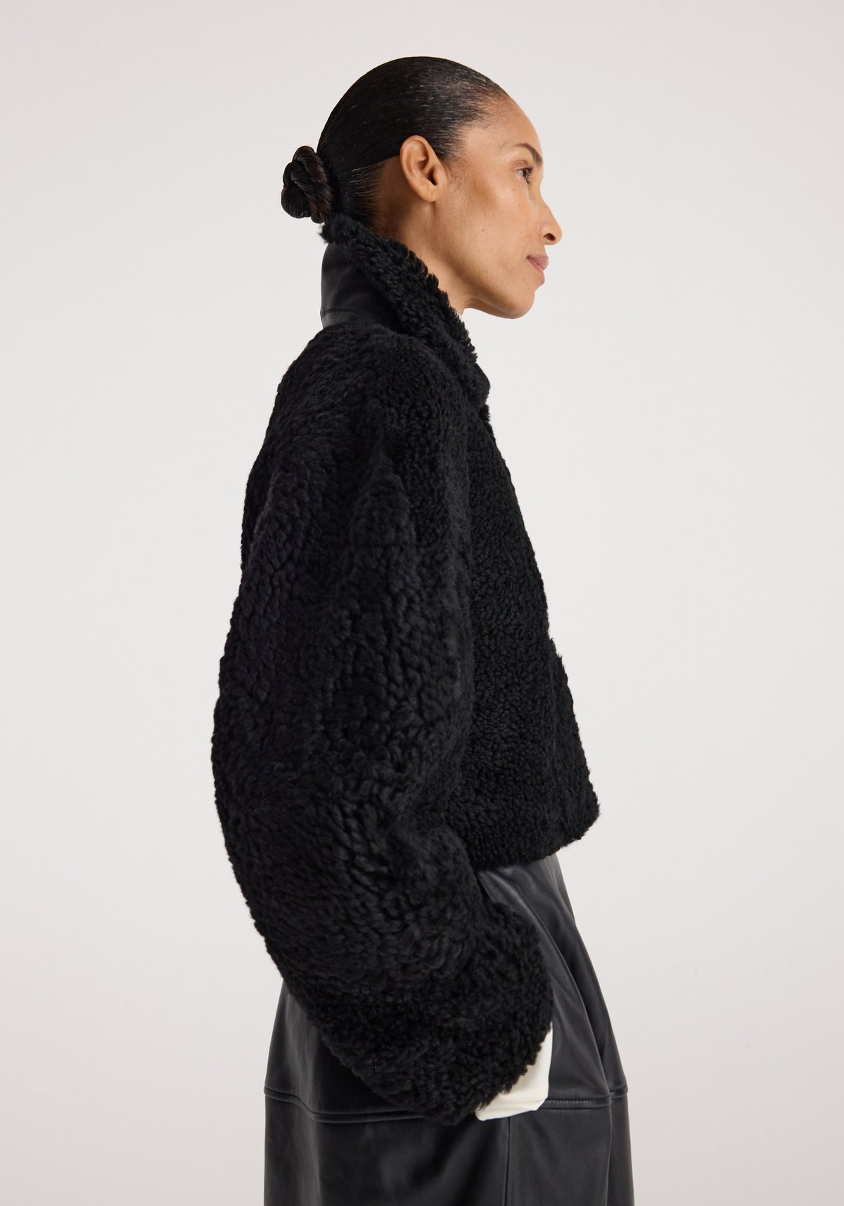 Woman wearing a black Rounded shearling jacket | black with a textured, cozy finish and high collar, paired with a black skirt, shown in a side profile against a plain background.