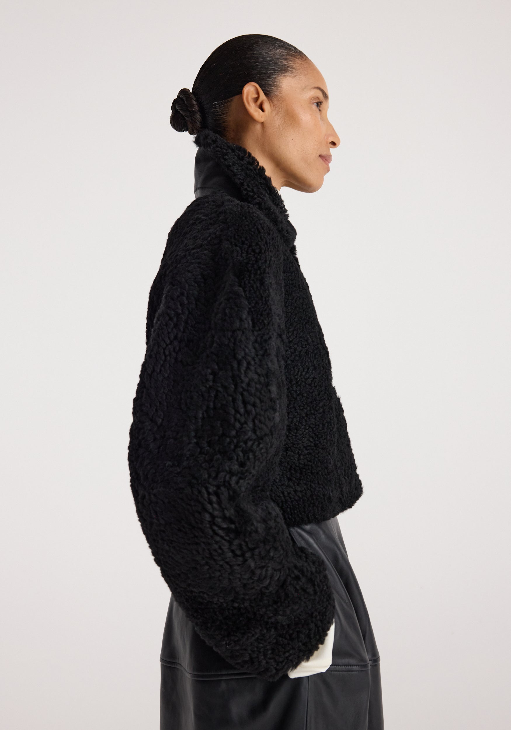 Woman wearing a black Rounded shearling jacket | black with a textured, cozy finish and high collar, paired with a black skirt, shown in a side profile against a plain background.