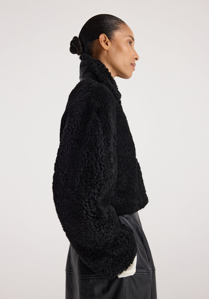 Woman wearing a black Rounded shearling jacket | black with a textured, cozy finish and high collar, paired with a black skirt, shown in a side profile against a plain background.
