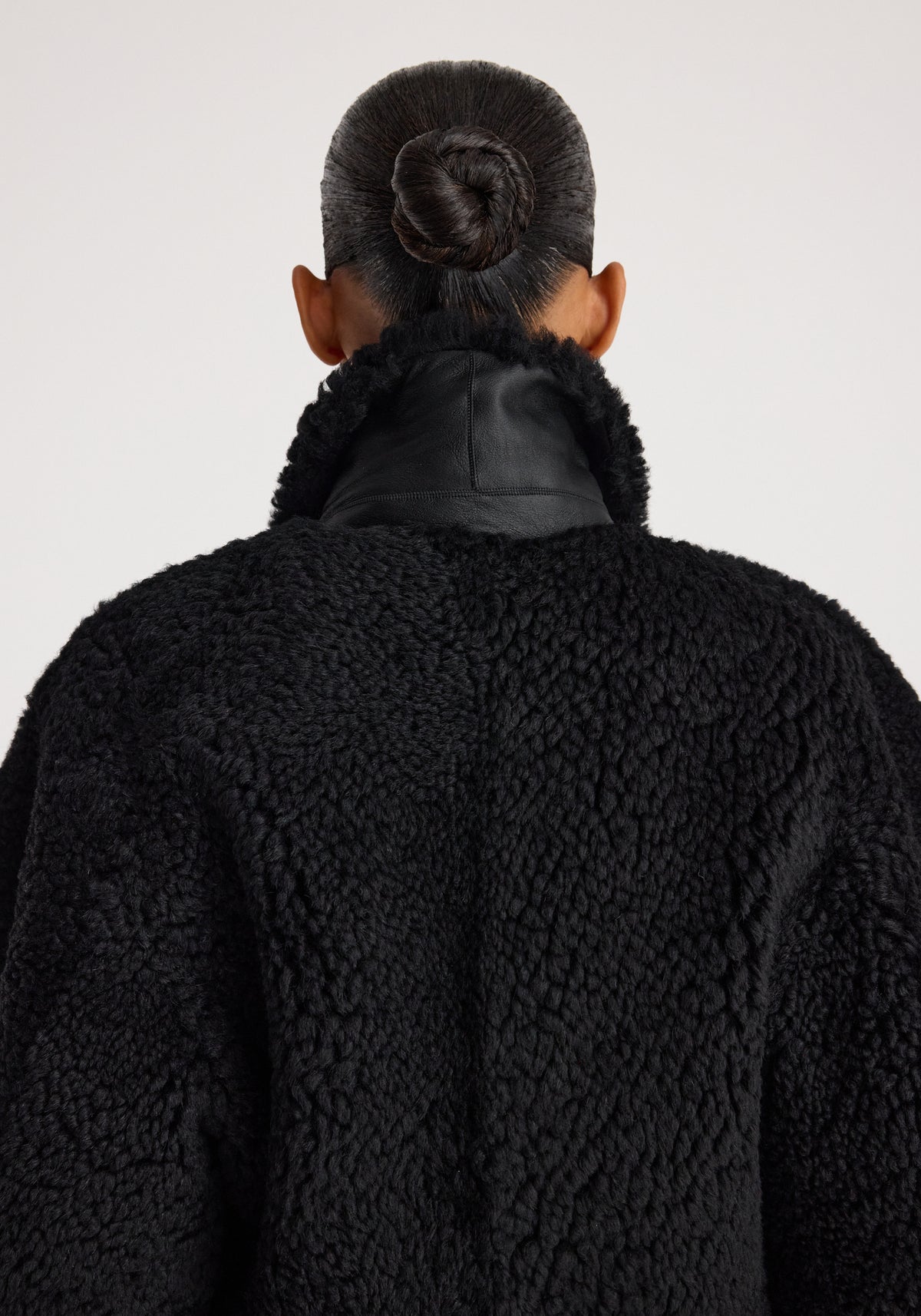 Back view of a person wearing a cozy Rounded shearling jacket | black with a textured, fluffy finish and a high collar, their hair styled in a neat bun.