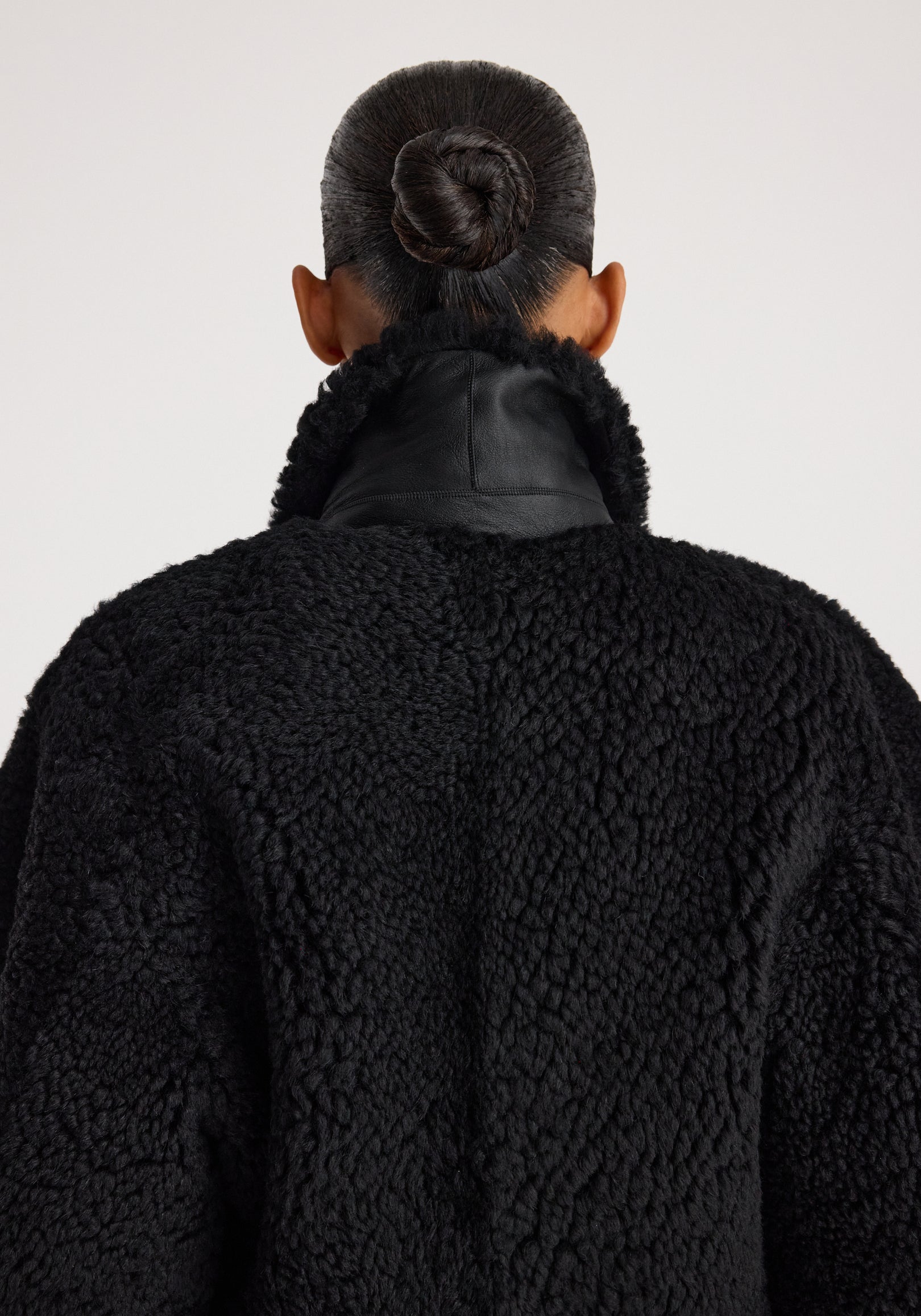 Back view of a person wearing a cozy Rounded shearling jacket | black with a textured, fluffy finish and a high collar, their hair styled in a neat bun.