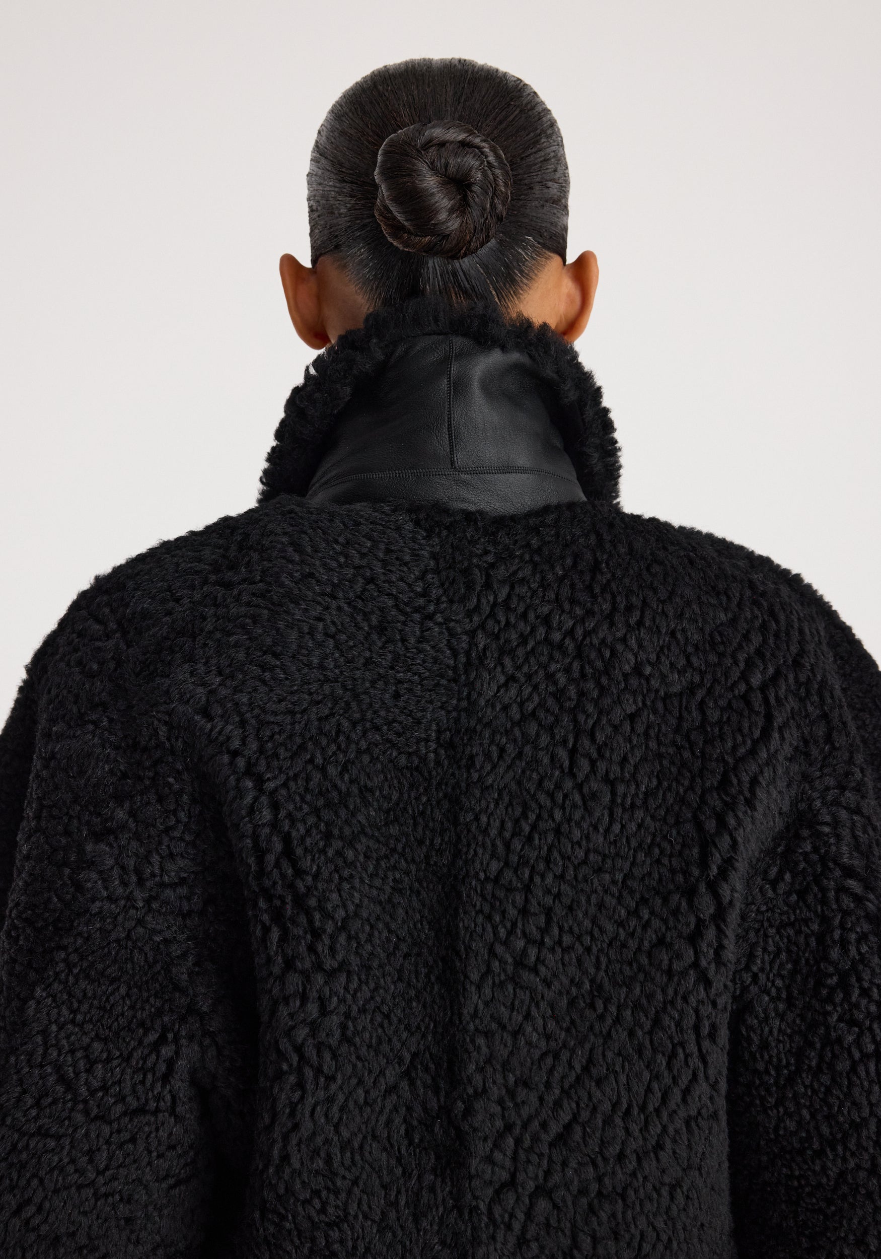 Back view of a person wearing a cozy Rounded shearling jacket | black with a textured, fluffy finish and a high collar, their hair styled in a neat bun.