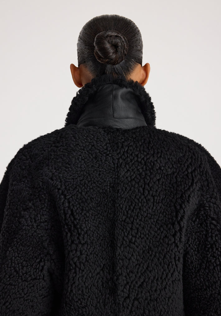 Back view of a person wearing a cozy Rounded shearling jacket | black with a textured, fluffy finish and a high collar, their hair styled in a neat bun.