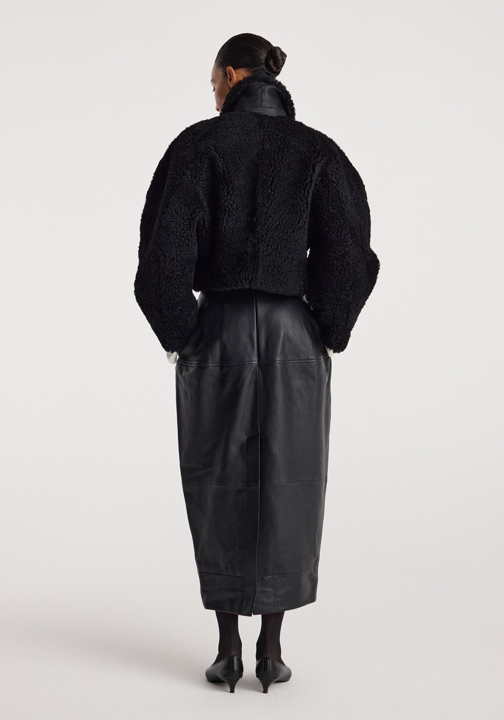 Woman wearing a black rounded shearling jacket | black with a high collar, paired with a long black leather skirt and black heels, standing with her back to the camera.