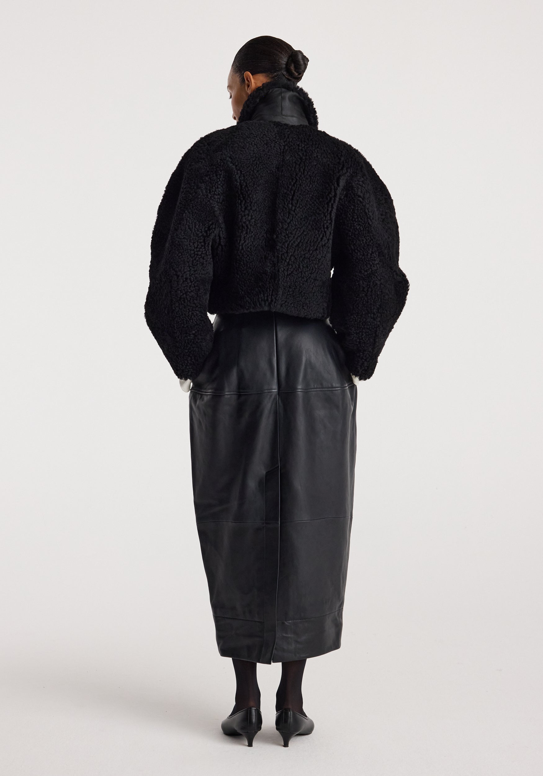 Woman wearing a black rounded shearling jacket | black with a high collar, paired with a long black leather skirt and black heels, standing with her back to the camera.