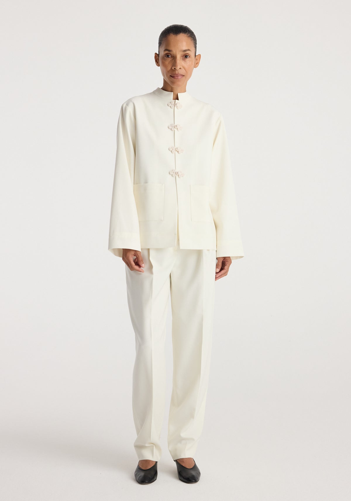 Woman wearing a minimalist off-white Pankou closure jacket | off white with long sleeves and front knot closures, paired with matching trousers and black flats.
