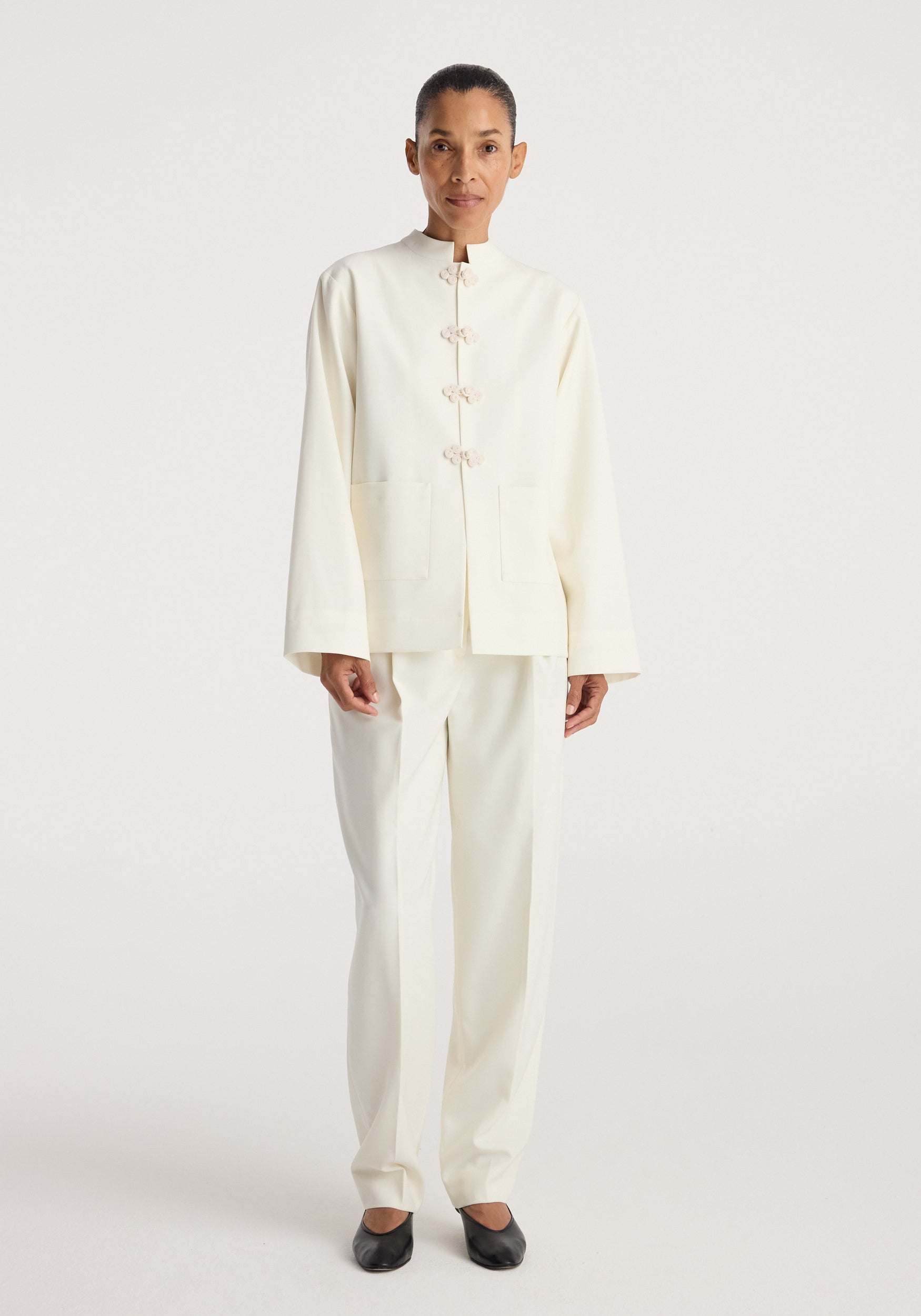 Woman wearing a minimalist off-white Pankou closure jacket | off white with long sleeves and front knot closures, paired with matching trousers and black flats.