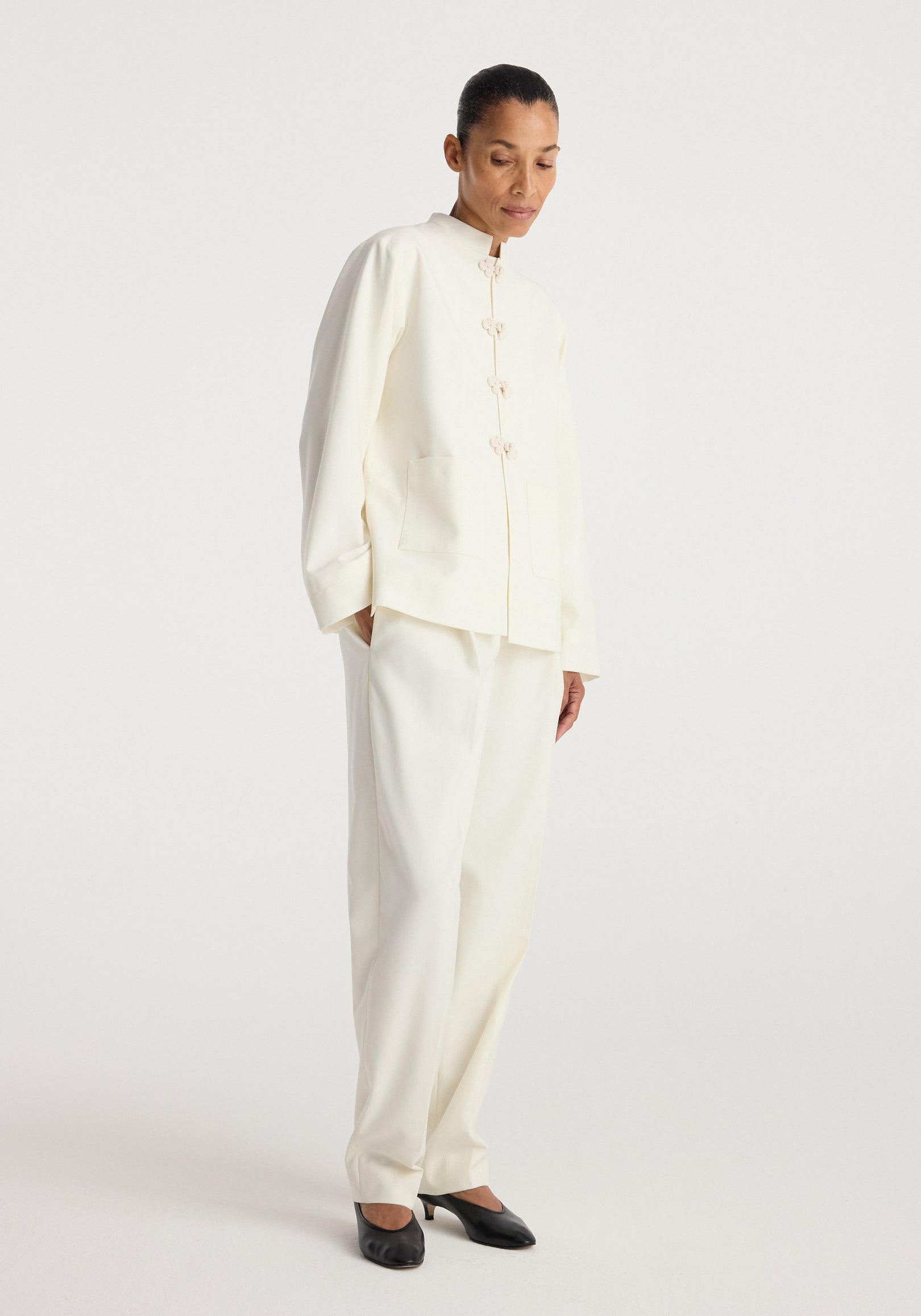Woman wearing an off-white Pankou closure jacket | off white with matching trousers and black heels, standing against a plain light background.