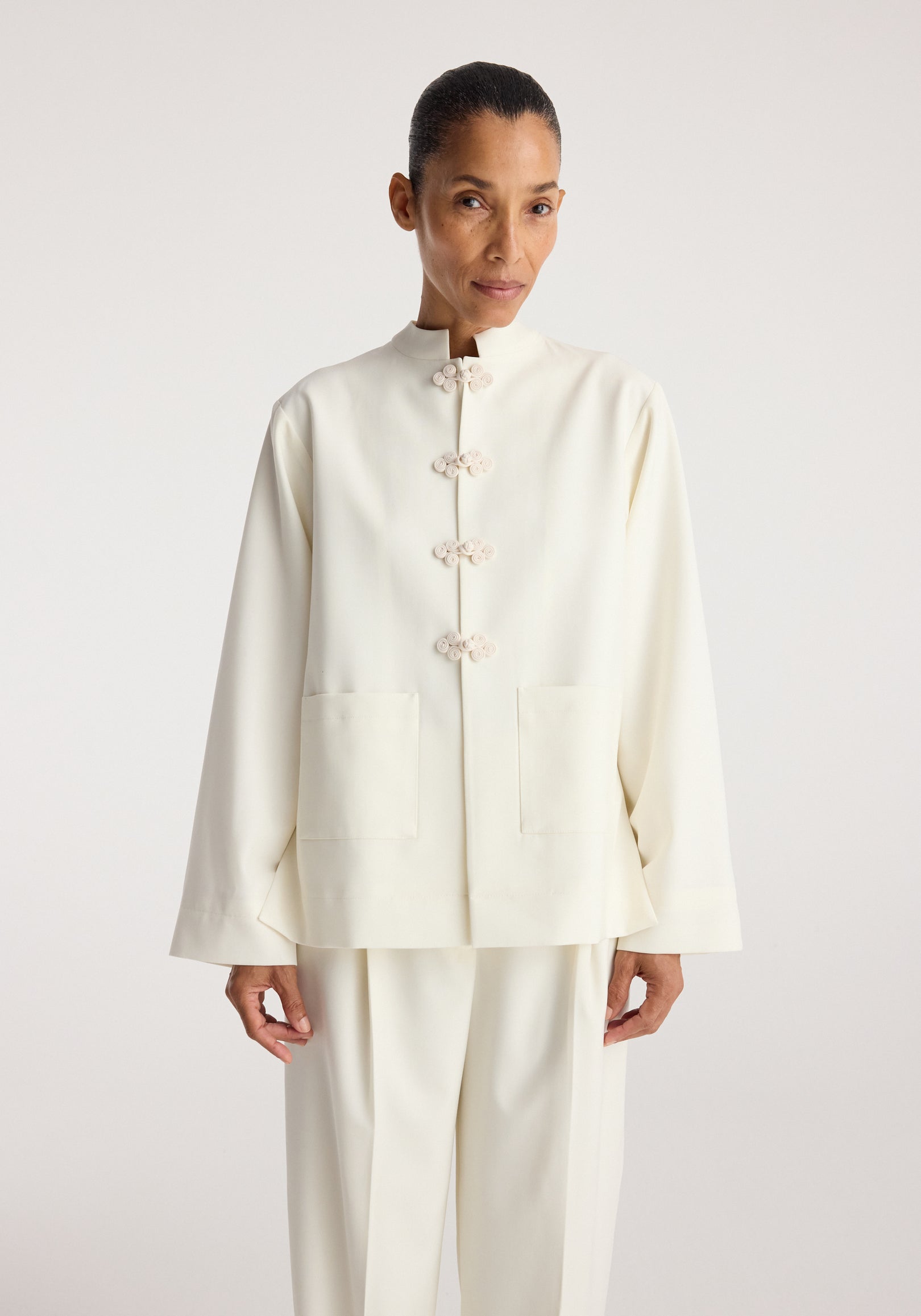 Woman wearing an off white Pankou closure jacket | off white with traditional frog button details, a stand collar, long sleeves, and front patch pockets.