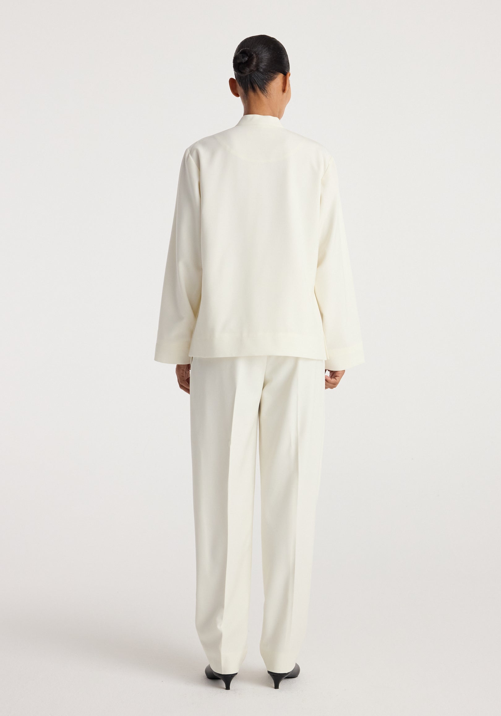 Back view of a person wearing a sleek, minimalist Pankou closure jacket | off white paired with matching tailored trousers and black pointed heels against a plain backdrop.