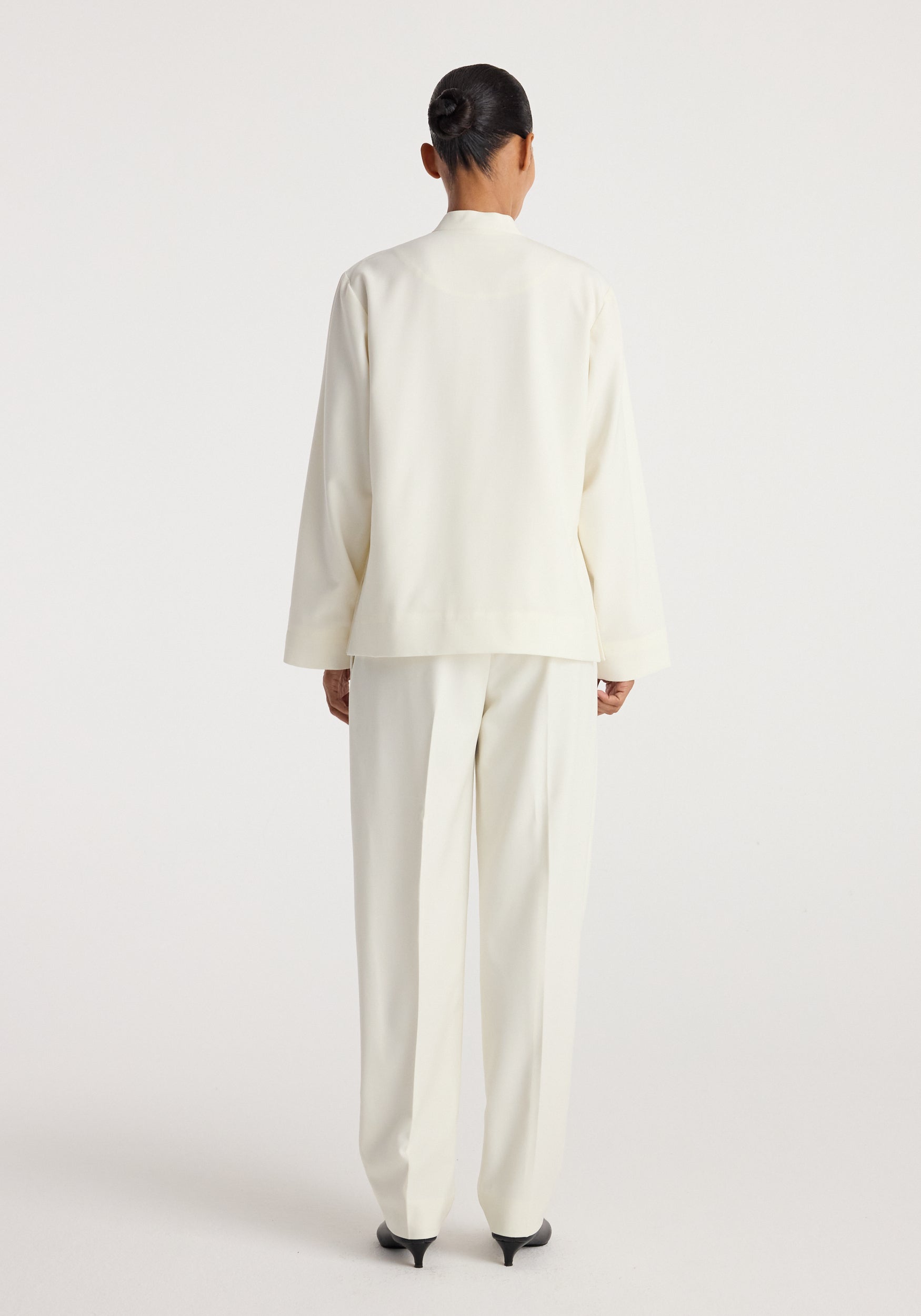 Back view of a person wearing a sleek, minimalist Pankou closure jacket | off white paired with matching tailored trousers and black pointed heels against a plain backdrop.