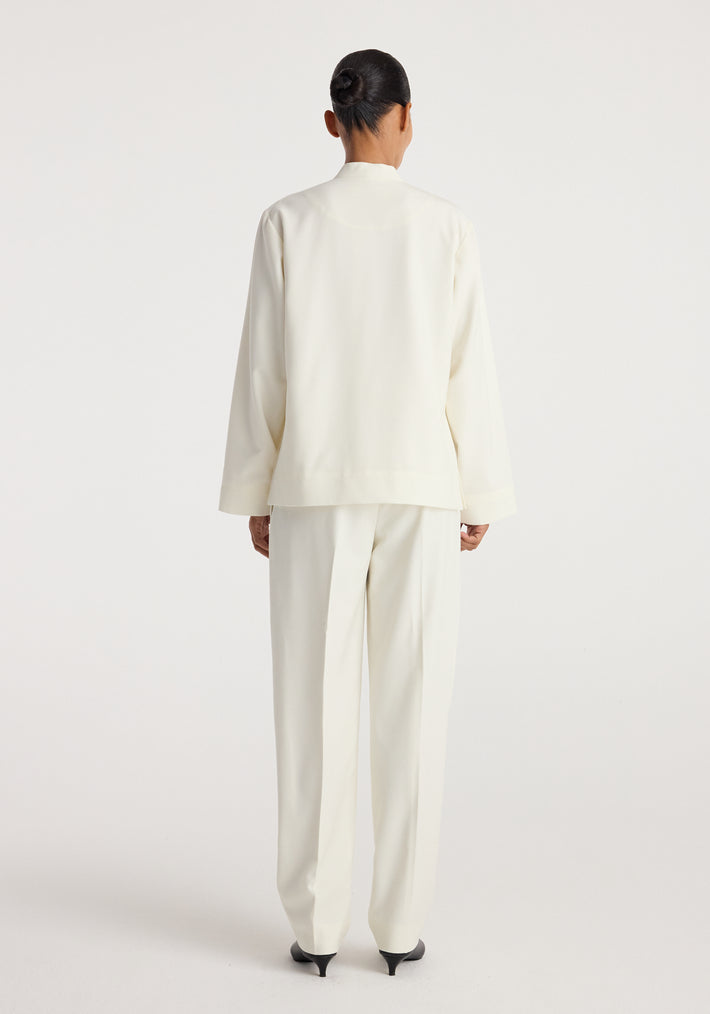 Back view of a person wearing a sleek, minimalist Pankou closure jacket | off white paired with matching tailored trousers and black pointed heels against a plain backdrop.