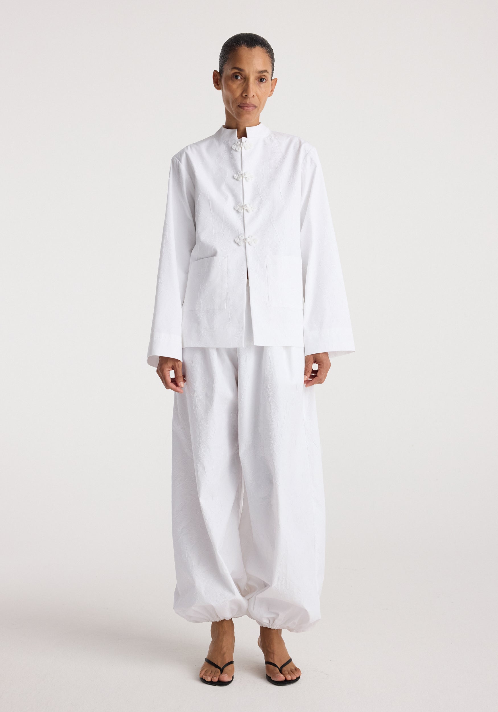 Woman wearing a minimalist white jacket with pankou closures paired with loose white pants and black sandals, styled on a plain light background, Pankou closure crushed jacket | optic white.