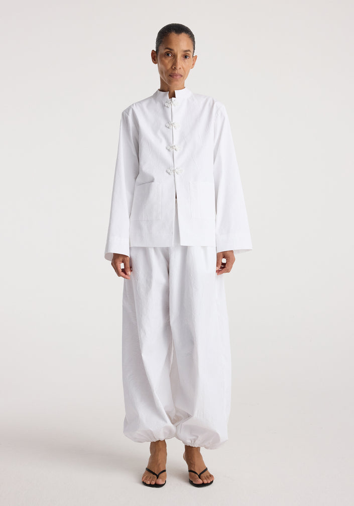 Woman wearing a minimalist white jacket with pankou closures paired with loose white pants and black sandals, styled on a plain light background, Pankou closure crushed jacket | optic white.