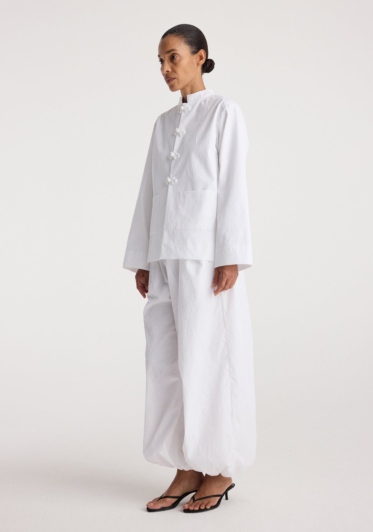 Woman wearing a white Pankou closure crushed jacket | optic white with long sleeves and matching loose pants, styled with simple black open-toe heels against a plain background.