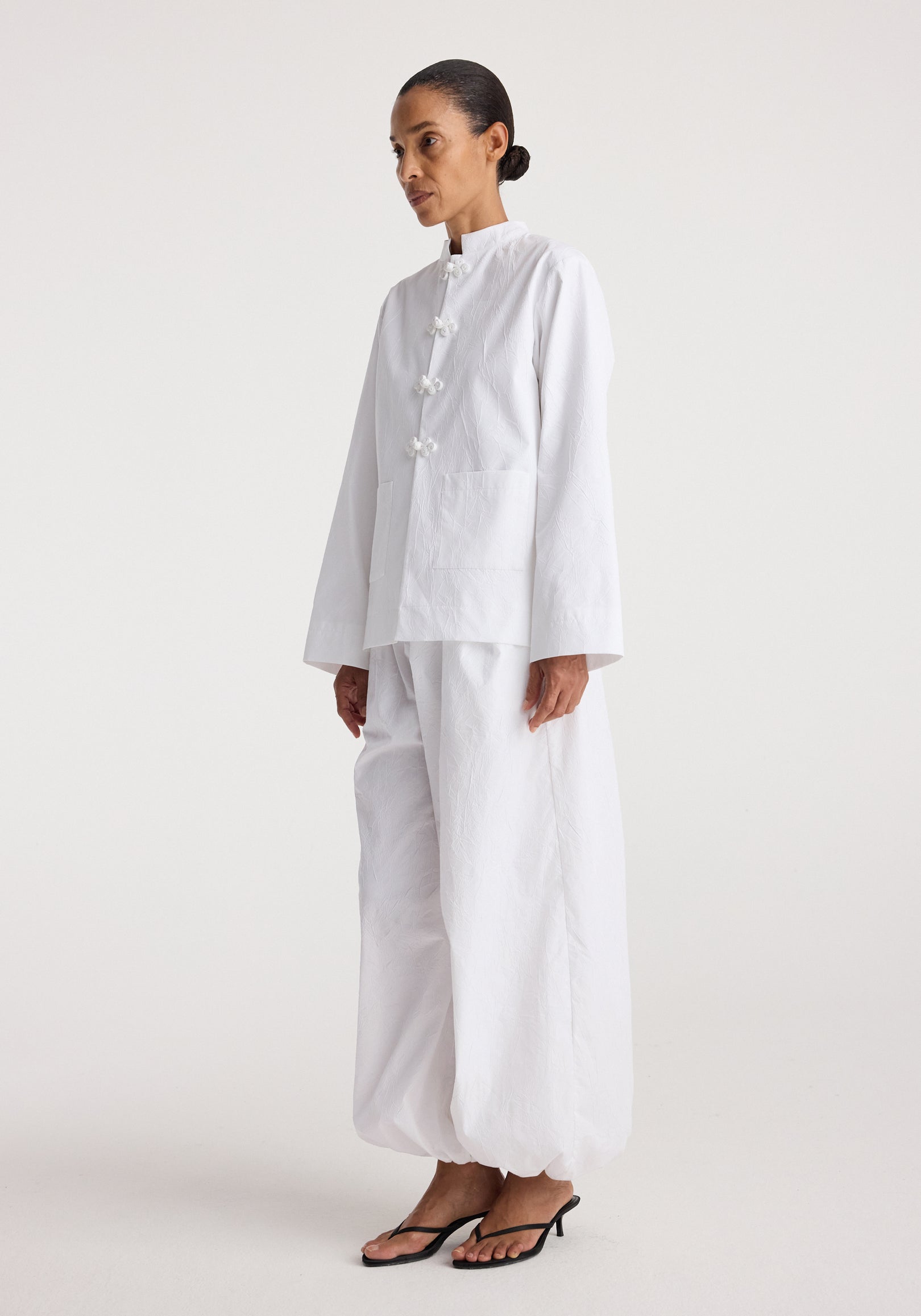 Woman wearing a white Pankou closure crushed jacket | optic white with long sleeves and matching loose pants, styled with simple black open-toe heels against a plain background.