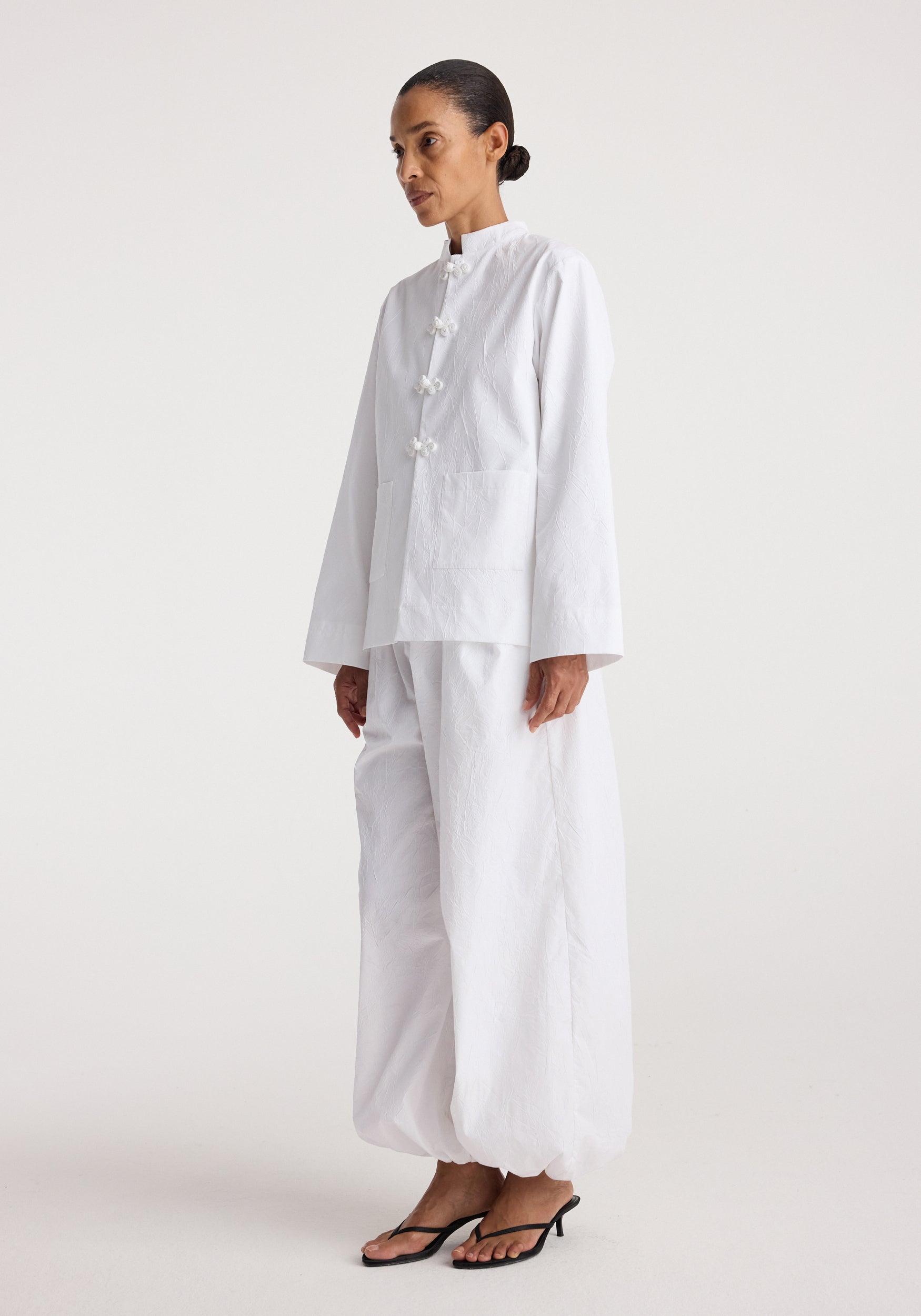 Woman wearing a white Pankou closure crushed jacket | optic white with long sleeves and matching loose pants, styled with simple black open-toe heels against a plain background.