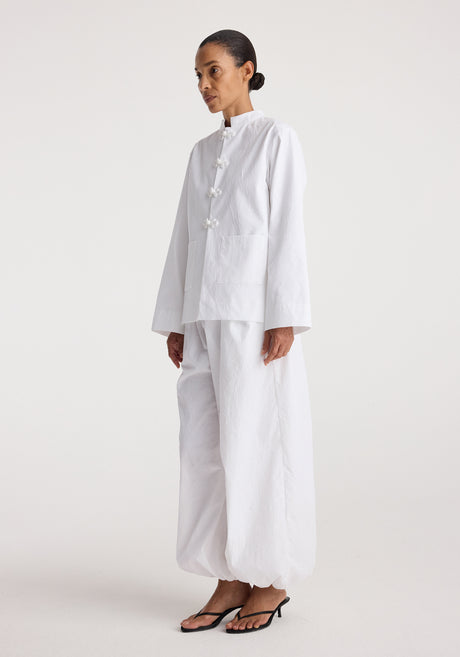 Woman wearing a minimalist white jacket with pankou closures paired with loose white pants and black sandals, styled on a plain light background, Pankou closure crushed jacket | optic white.