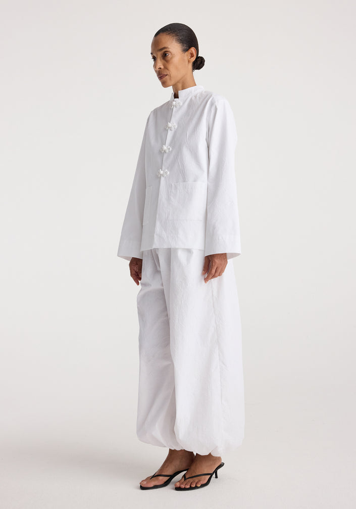 Woman wearing a minimalist white jacket with pankou closures paired with loose white pants and black sandals, styled on a plain light background, Pankou closure crushed jacket | optic white.