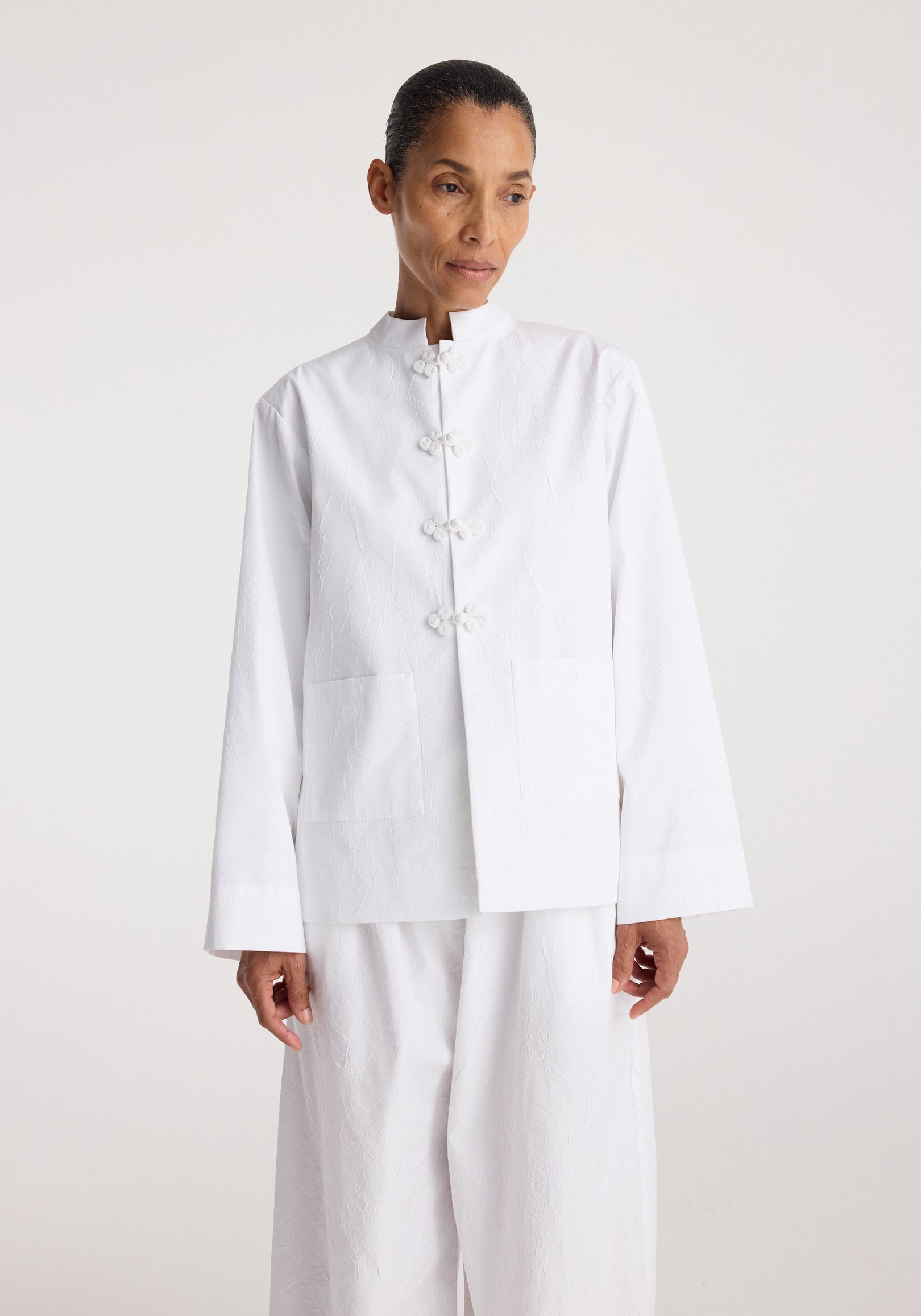 Woman wearing a Pankou closure crushed jacket | optic white with traditional Chinese knot buttons, featuring a high collar and large front pockets, styled with matching white pants.