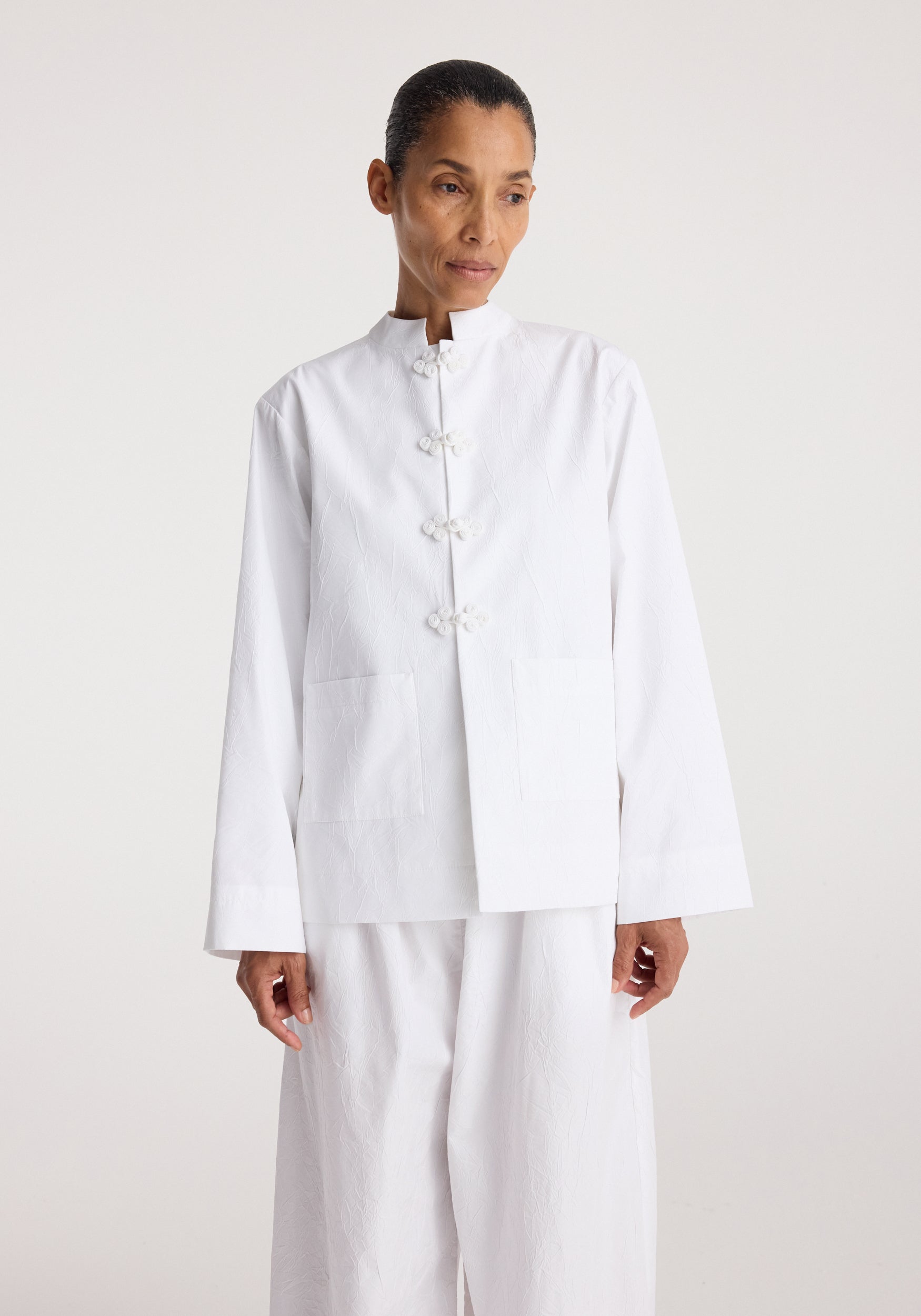 Woman wearing a Pankou closure crushed jacket | optic white with traditional Chinese knot buttons, featuring a high collar and large front pockets, styled with matching white pants.