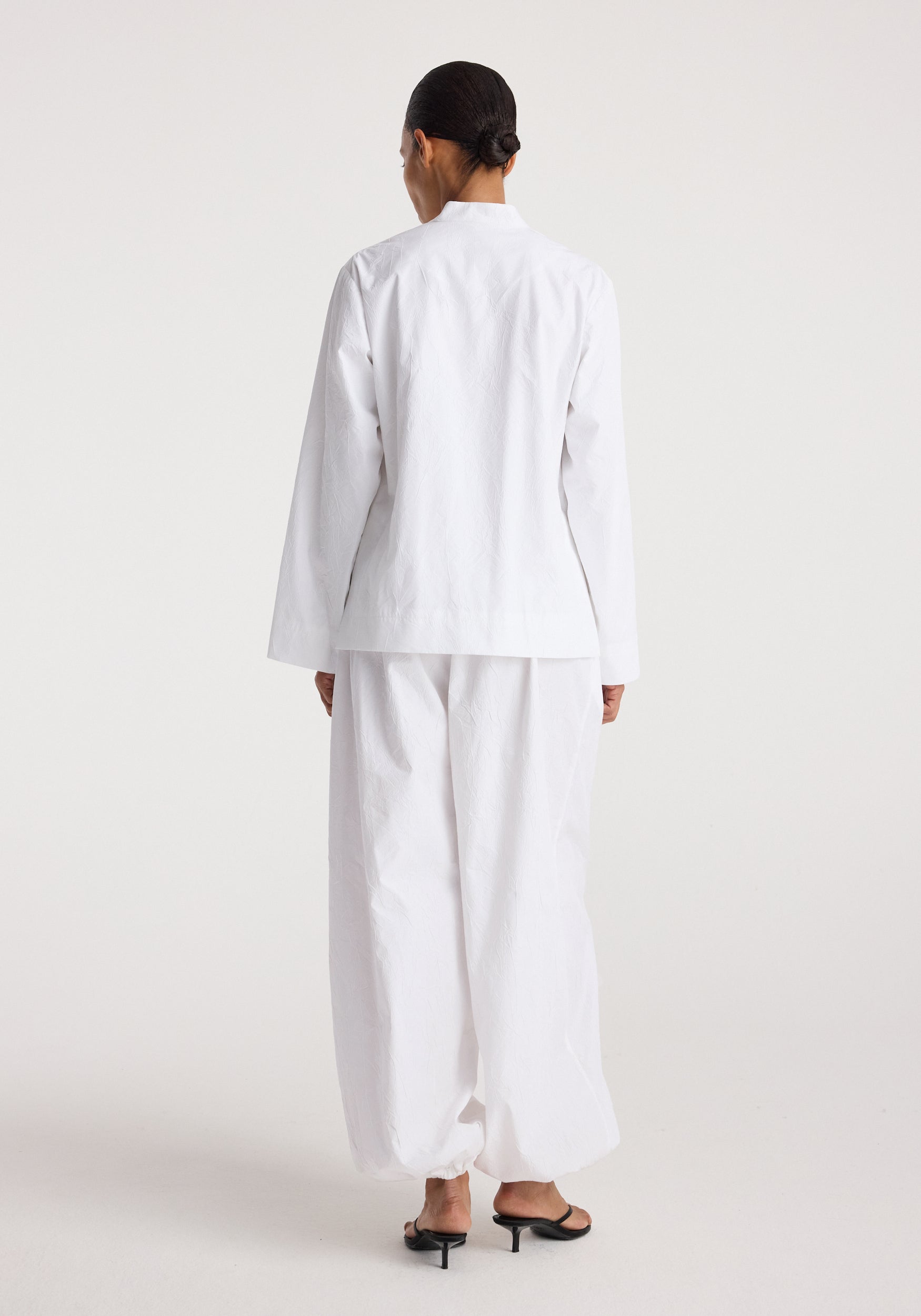 Woman wearing Pankou closure crushed jacket | optic white, shown from the back with loose white pants and black open-toe heels against a plain background.