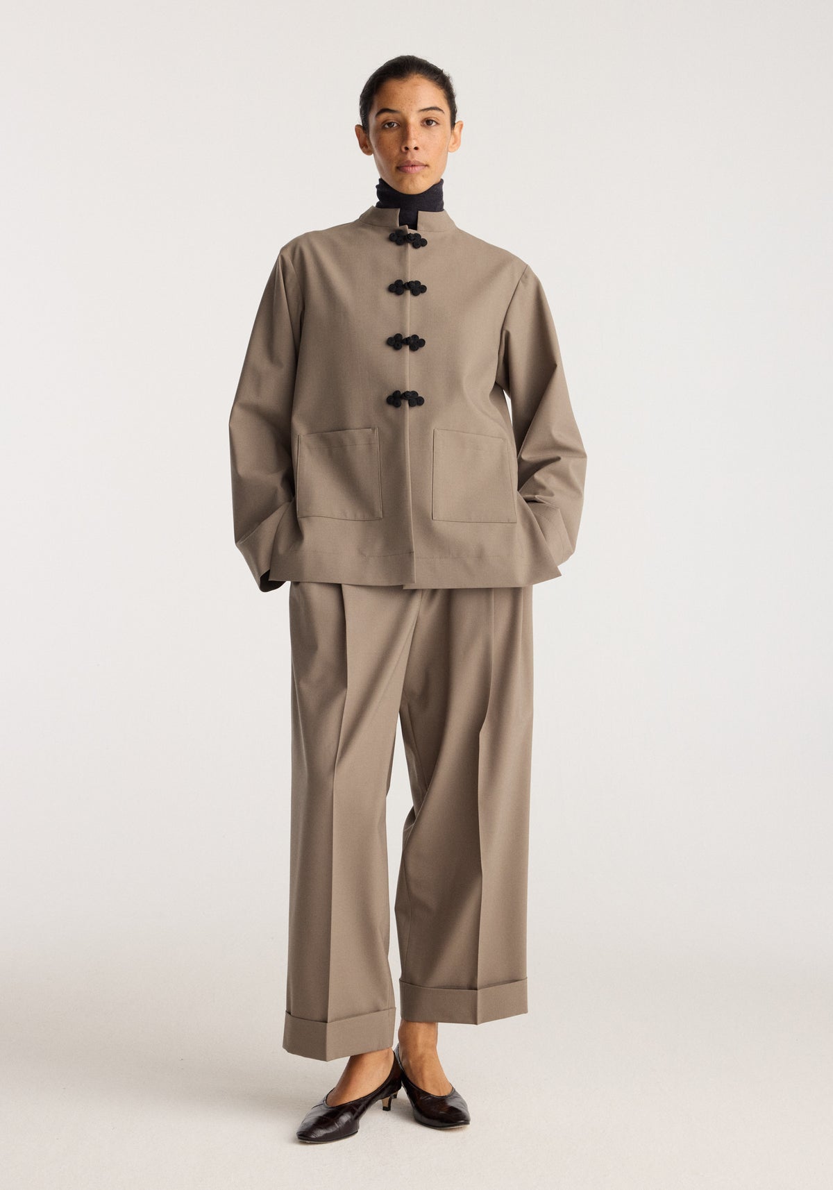 Pankou closure jacket | taupe worn by a model with black frog closures a high collar and relaxed tailoring styled with cropped taupe trousers and loafers