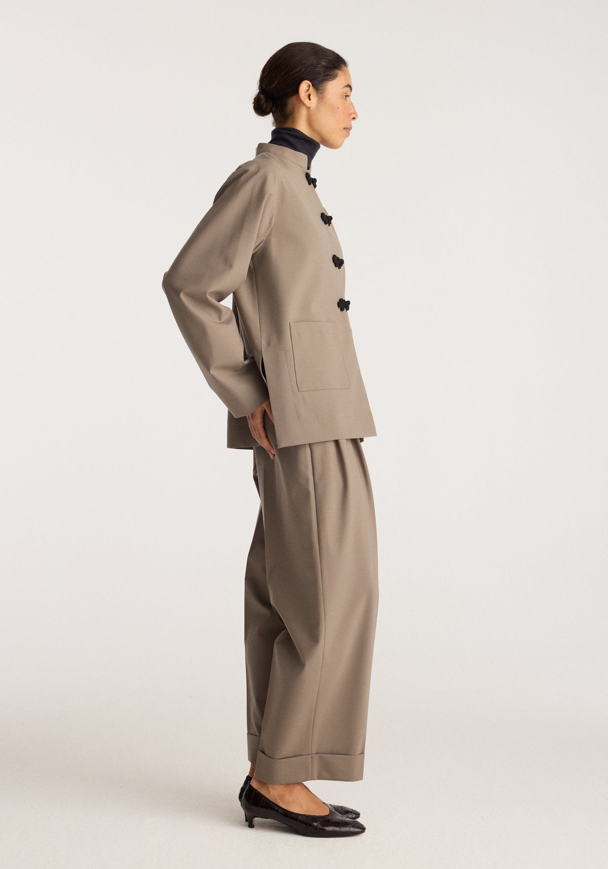 Pankou closure jacket | taupe shown in side profile with black knot closures high collar long sleeves and a tailored silhouette worn with taupe trousers