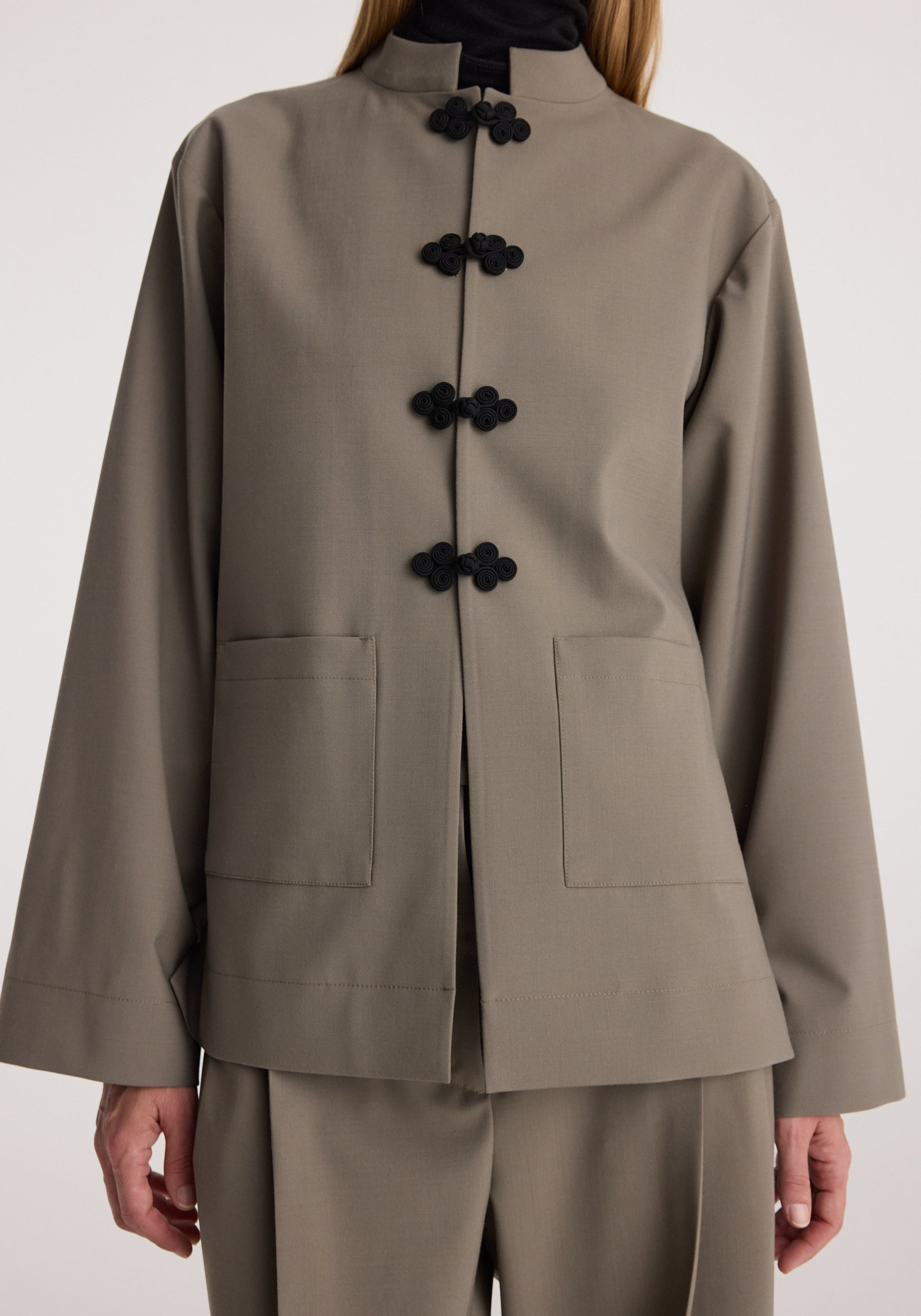 Close-up of a person wearing a taupe Pankou closure jacket | taupe with black traditional toggle buttons, front pockets, and a high collar over matching pants.