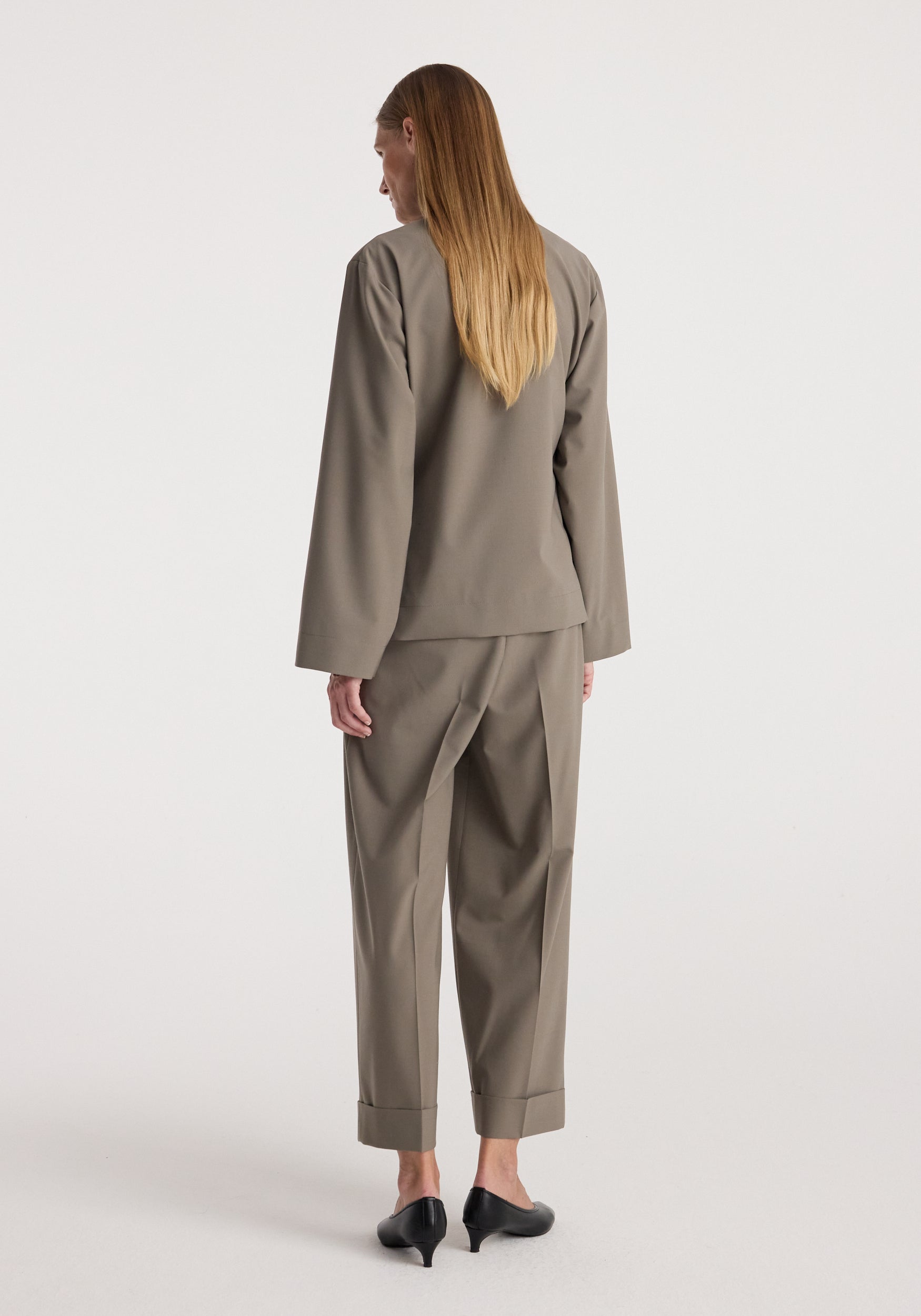 Woman with long straight hair wearing a taupe Pankou closure jacket | taupe and matching trousers, viewed from the back, standing on a plain background.