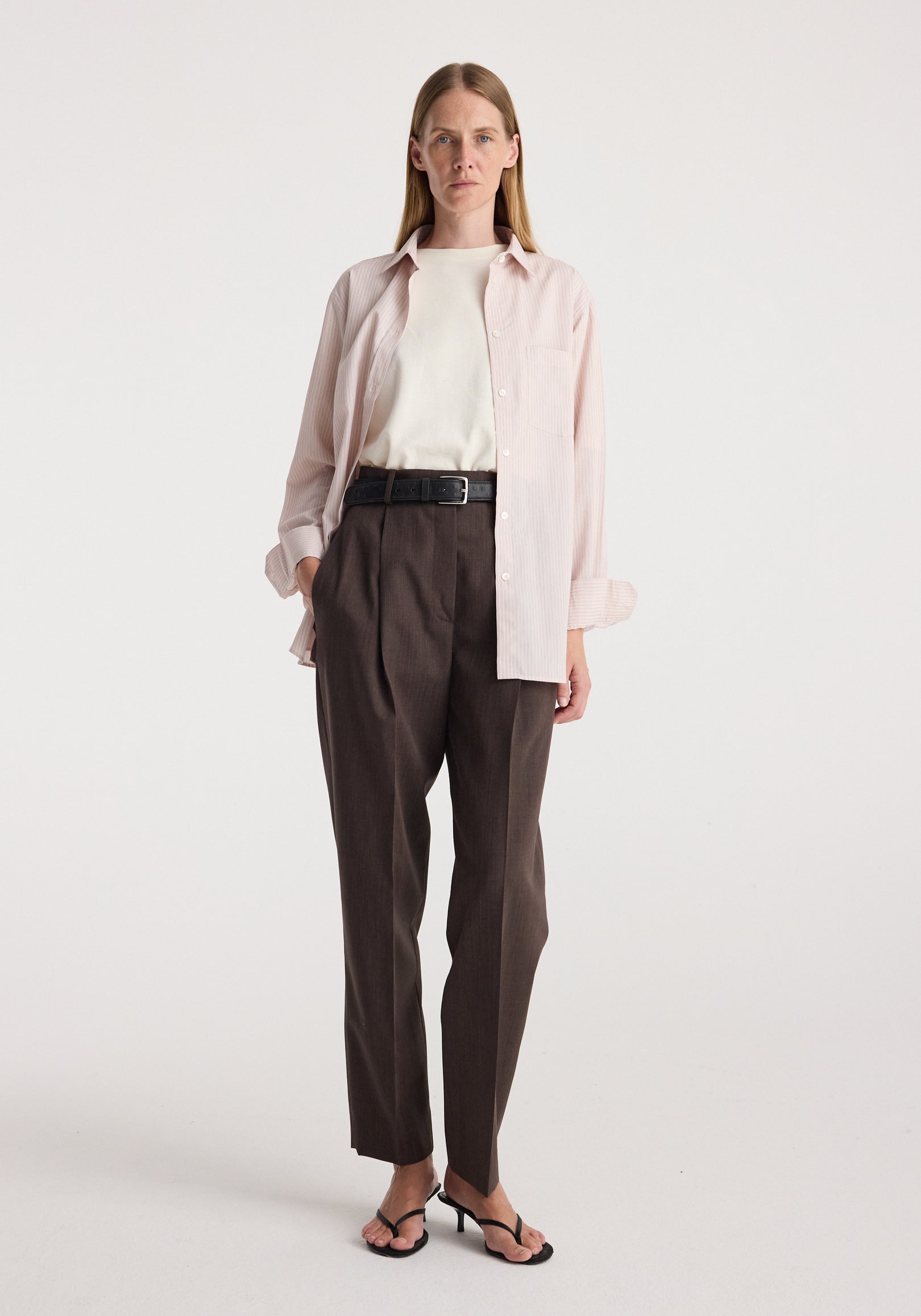 Woman wearing a Classic fit striped shirt | rose stripe in light pink over a white top, paired with high-waisted dark brown pants and black heeled sandals against a plain background.