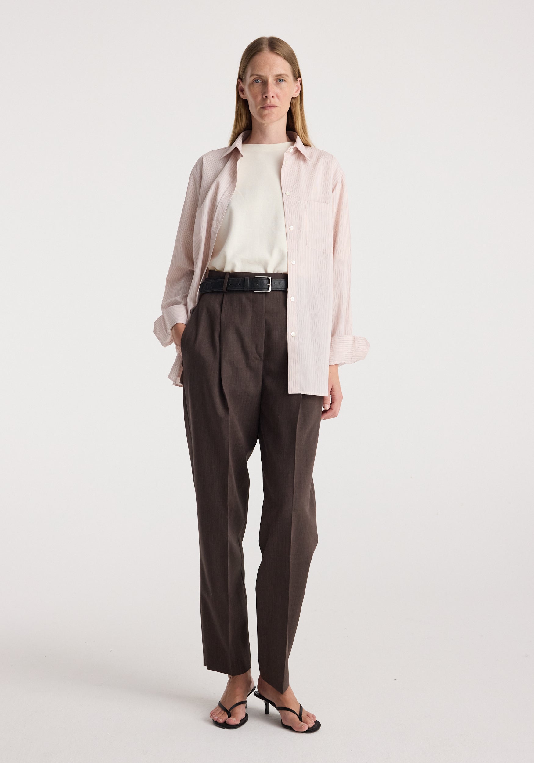 Woman wearing a Classic fit striped shirt | rose stripe in light pink over a white top, paired with high-waisted dark brown pants and black heeled sandals against a plain background.