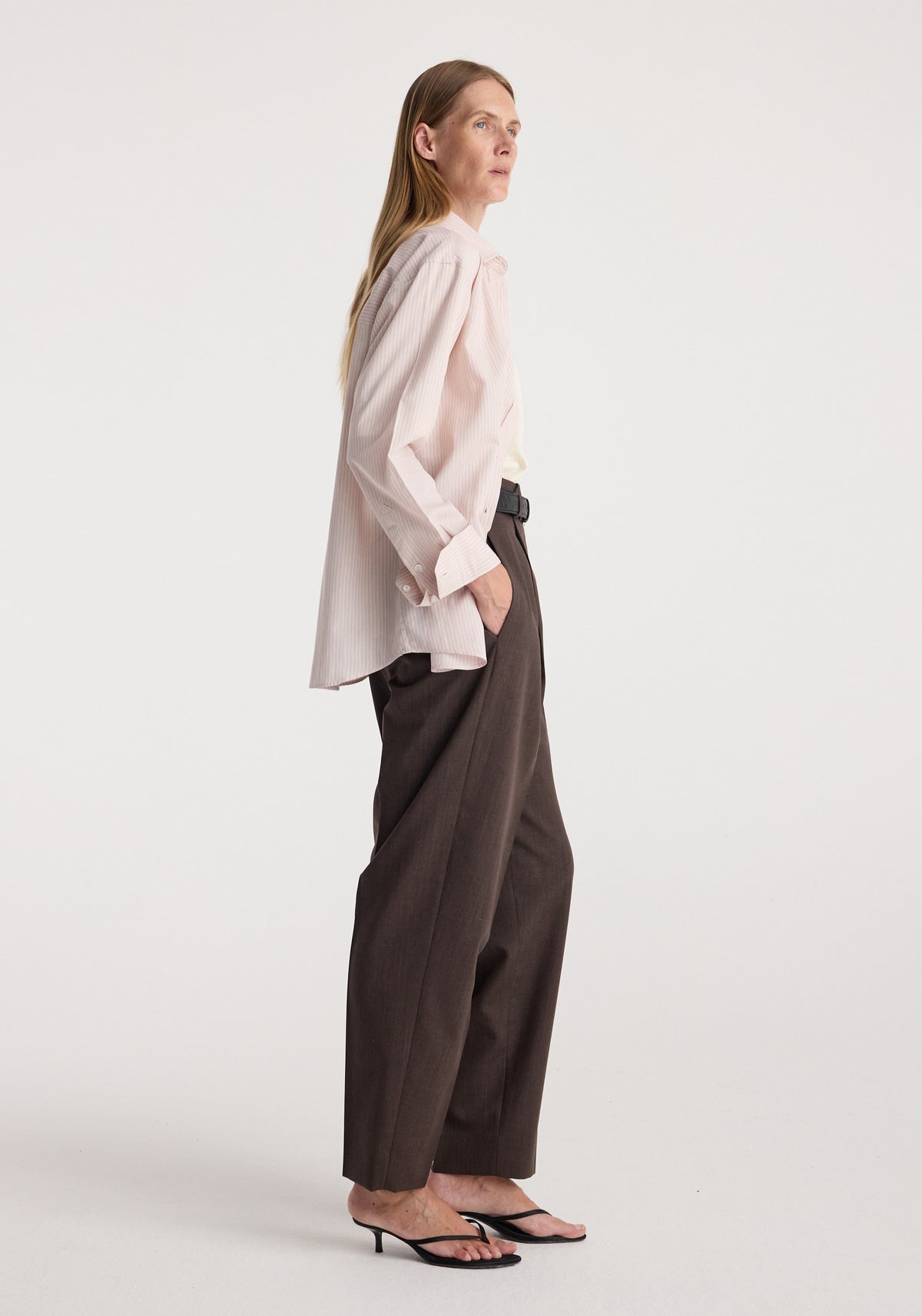 Woman wearing a Classic fit striped shirt | rose stripe styled with dark brown wide-leg trousers and black heeled sandals, standing in profile against a plain background.