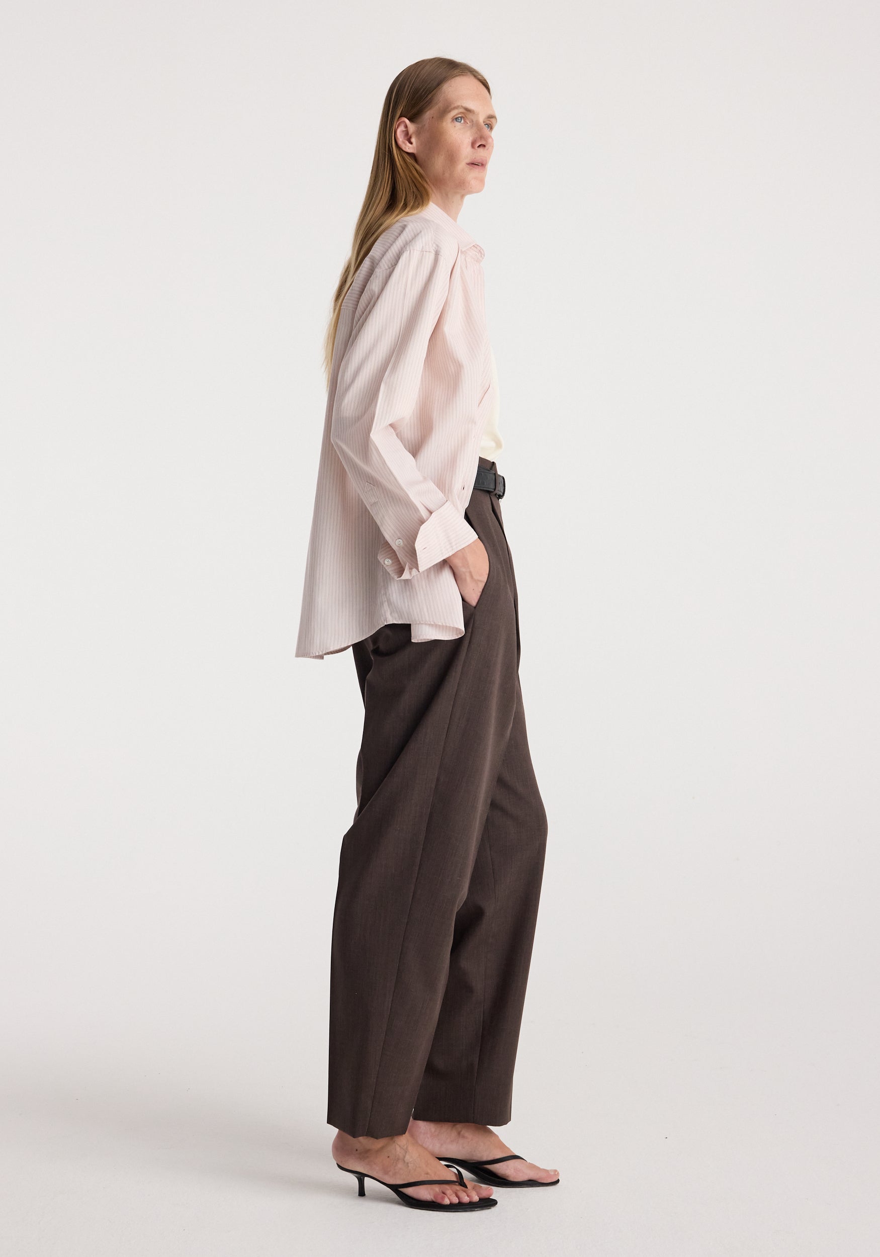Woman wearing a Classic fit striped shirt | rose stripe styled with dark brown wide-leg trousers and black heeled sandals, standing in profile against a plain background.