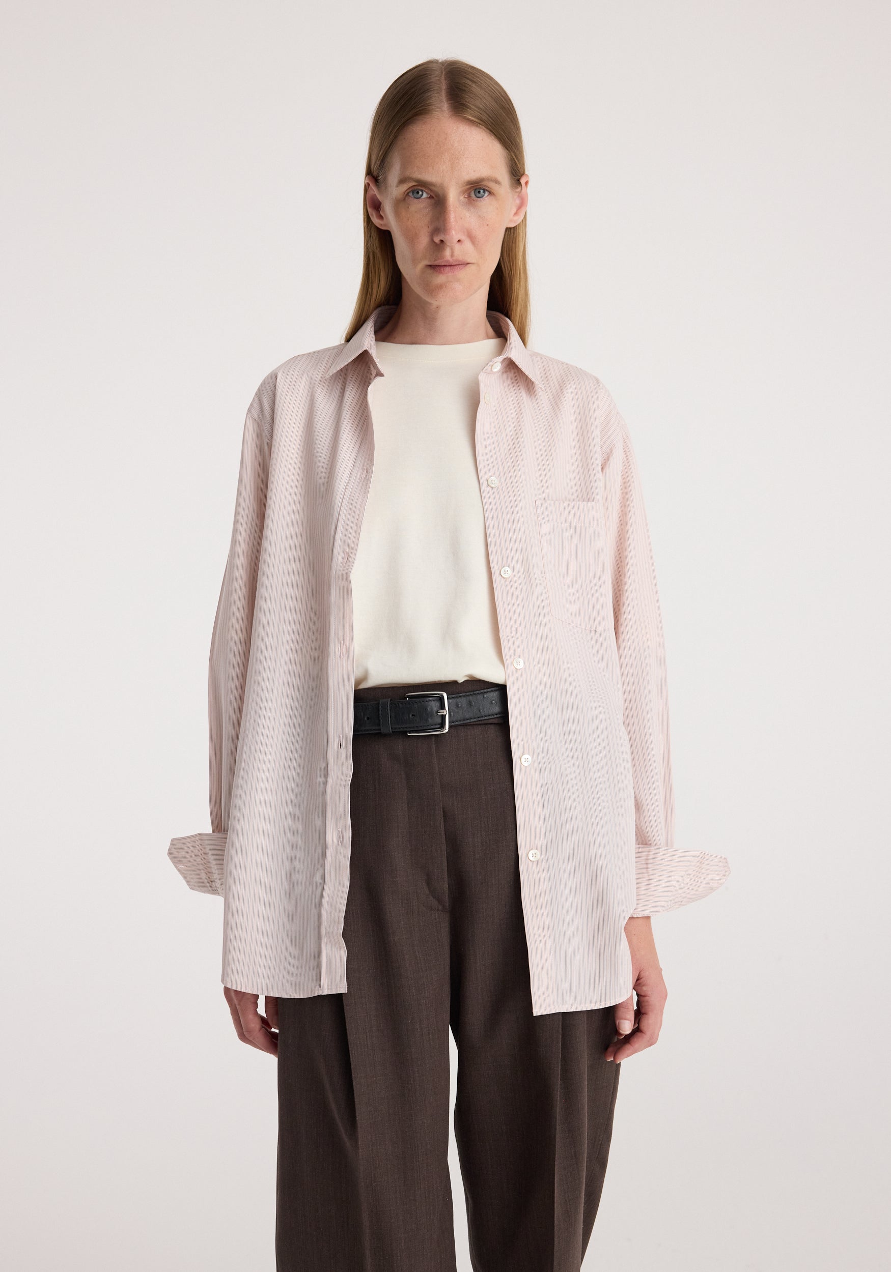 Woman wearing a Classic fit striped shirt | rose stripe, unbuttoned over a white top with brown high-waisted pants and a black belt in a neutral studio setting.