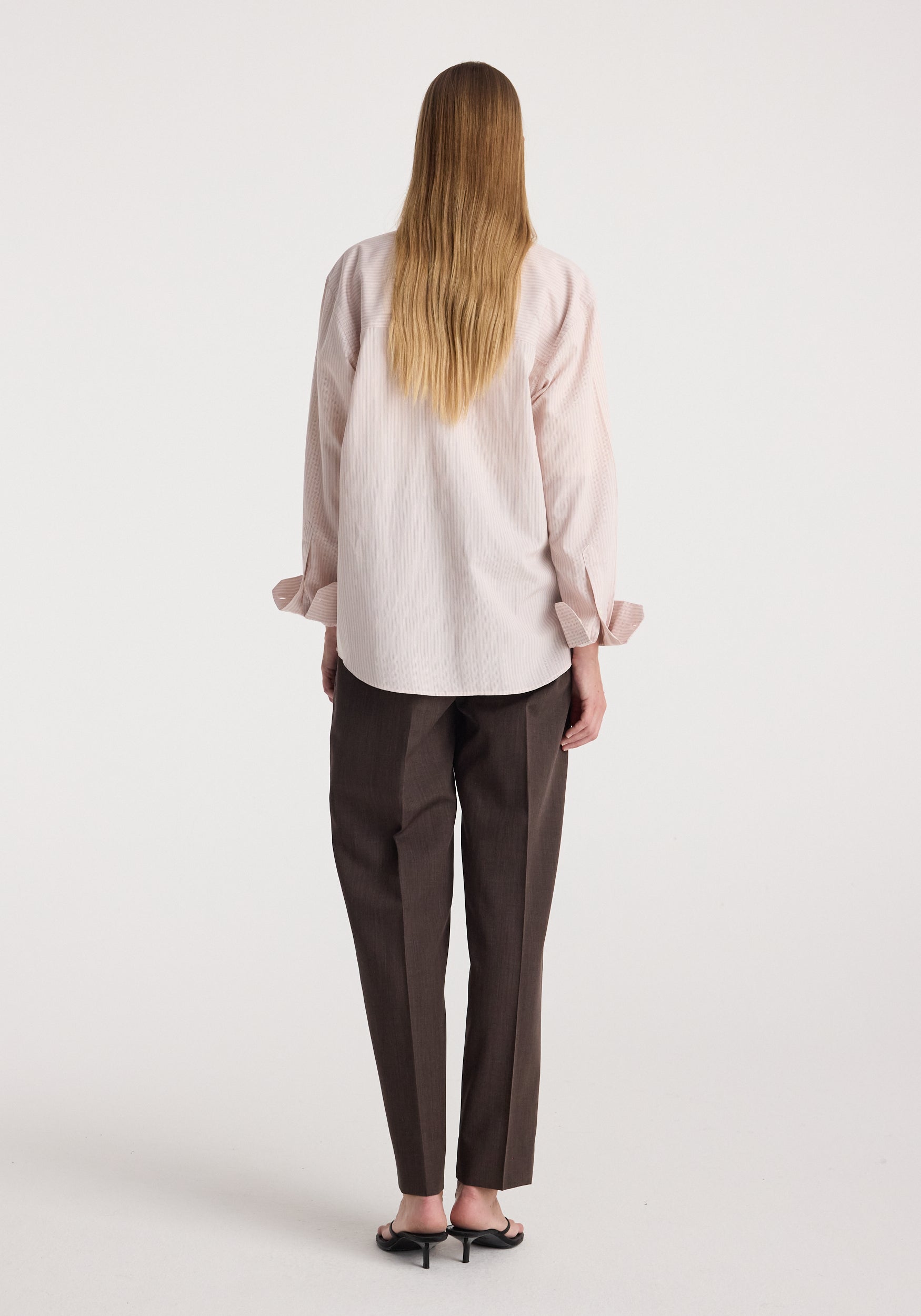 Woman seen from the back wearing a Classic fit striped shirt | rose stripe in light pink with long sleeves and dark brown tailored pants, standing against a plain background.