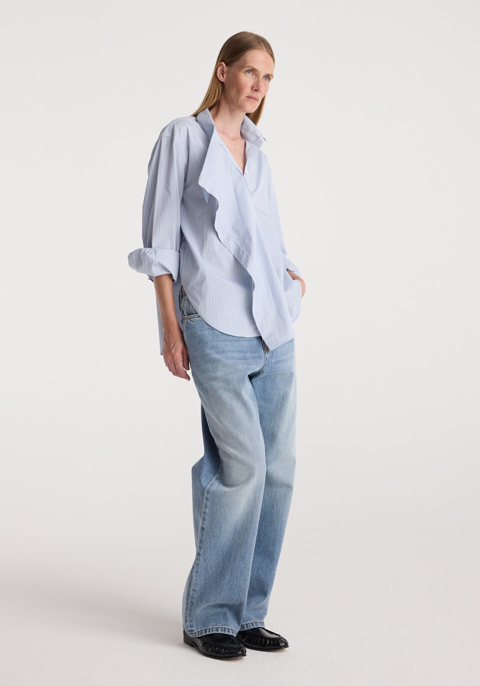 Woman wearing a light blue striped Draped collar shirt | blue stripe with rolled-up sleeves paired with loose-fitting light blue jeans and black shoes against a plain background.
